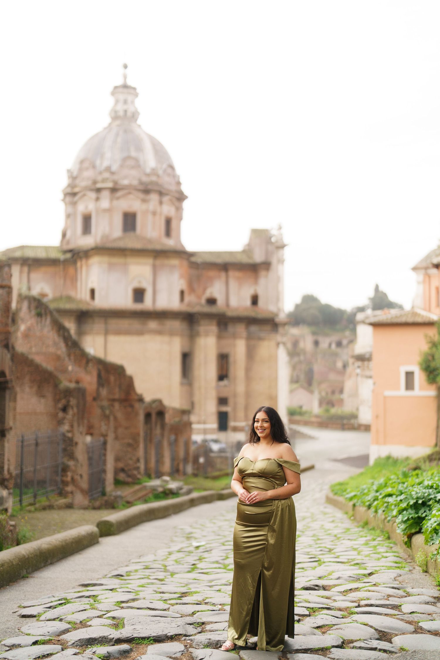 Pregnant woman in olive green gown poses on cobblestone path before Roman ruins and domed church in Rome.