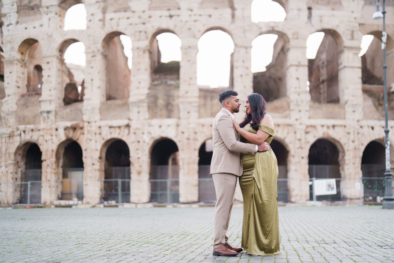 Couple embracing in front of the Colosseum in Rome, Italy, wearing elegant attire for a romantic engagement photo.