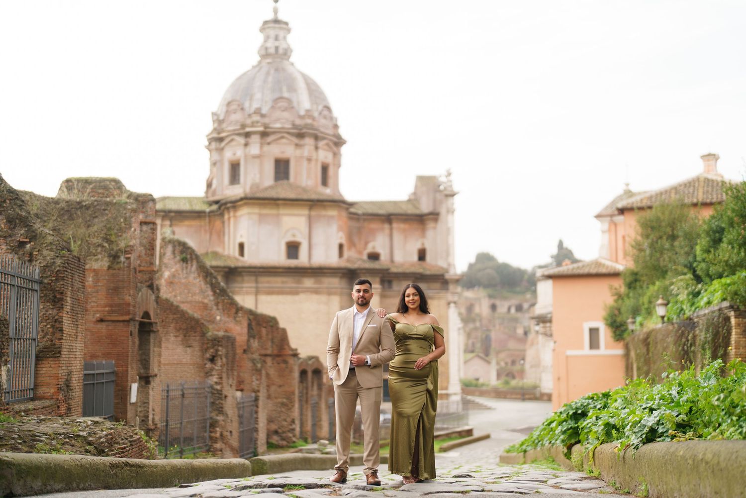 Couple posing in front of ancient Roman ruins and domed church on cobblestone path in Rome, Italy.