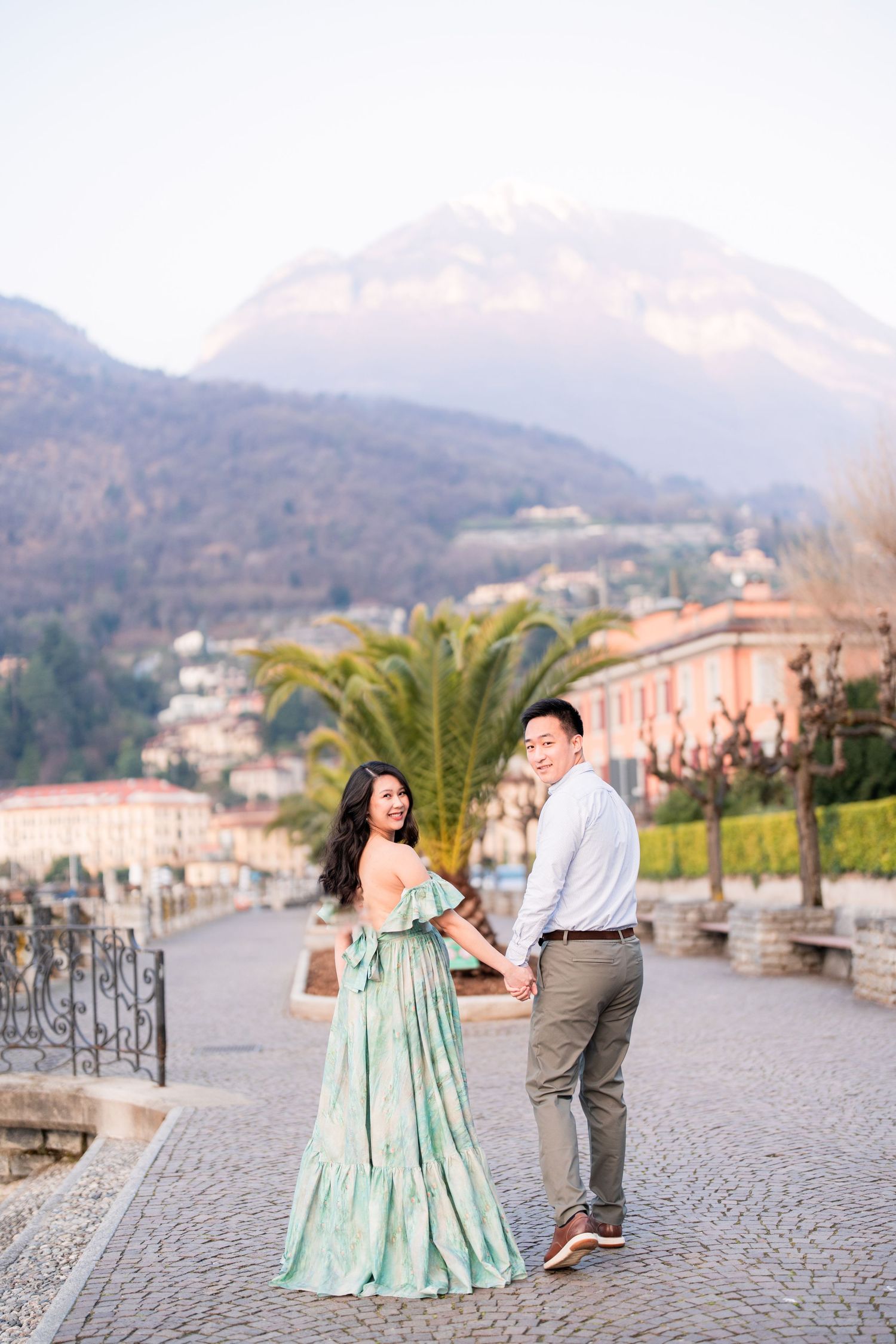 Couple holding hands on cobblestone path with mountain backdrop and palm trees in scenic Italian lakeside town.