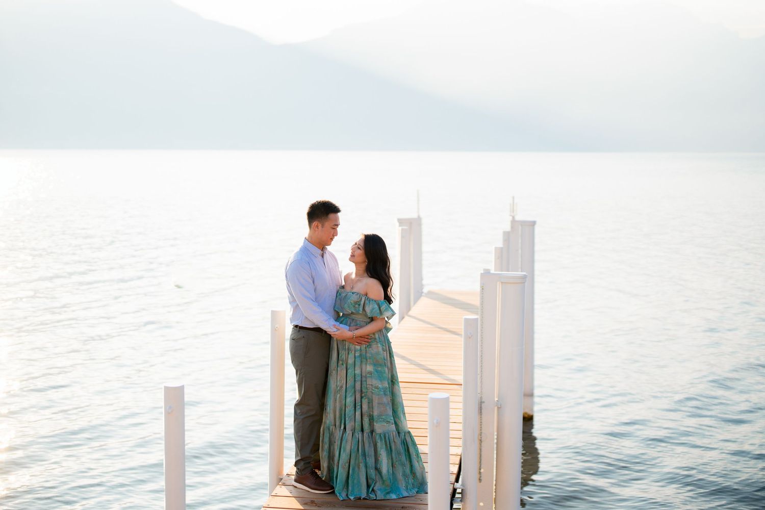 Couple embracing on a lakeside dock with misty mountains and calm blue water in the background.