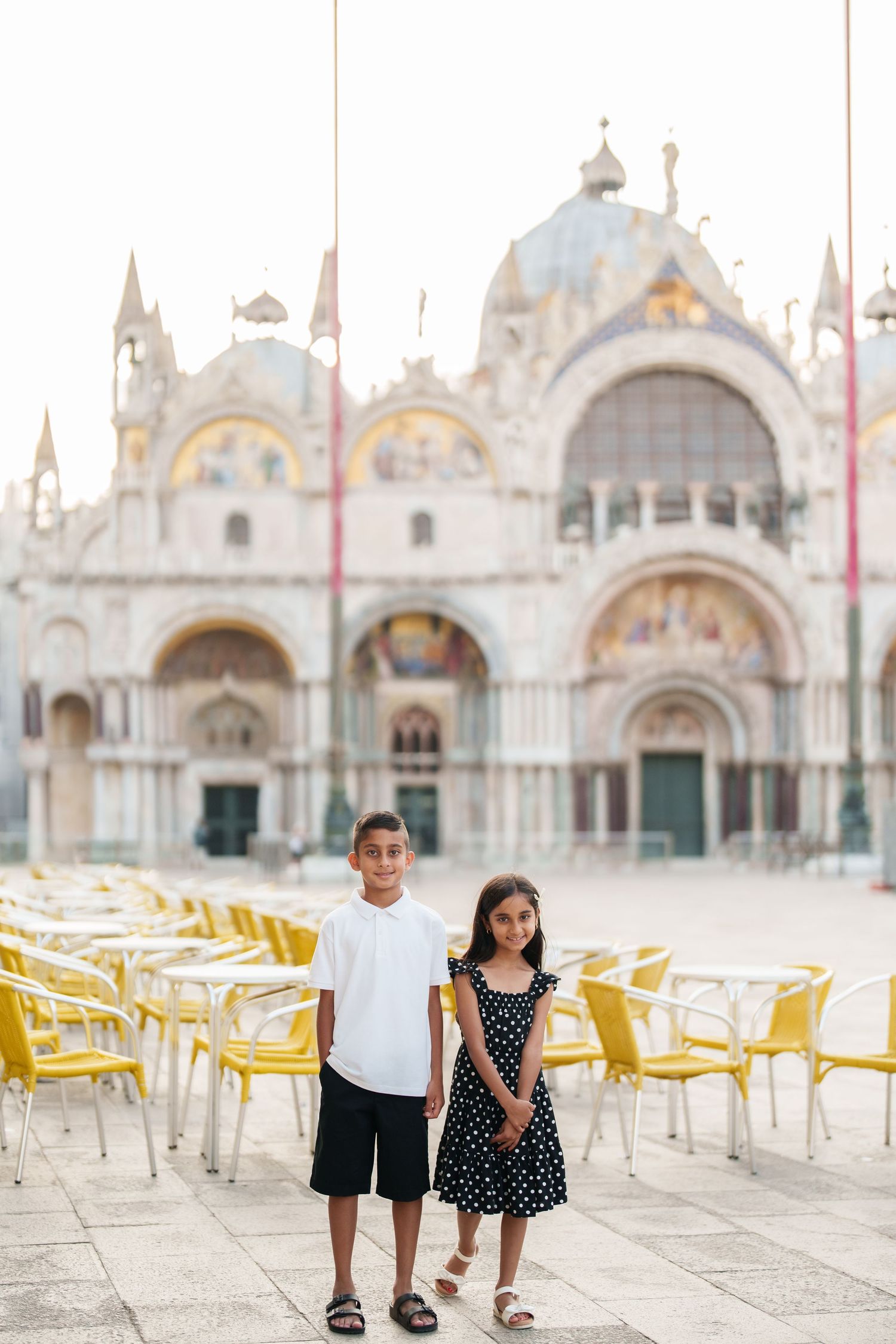 Two children posing in St. Mark's Square, Venice, with the iconic Basilica di San Marco in the background.