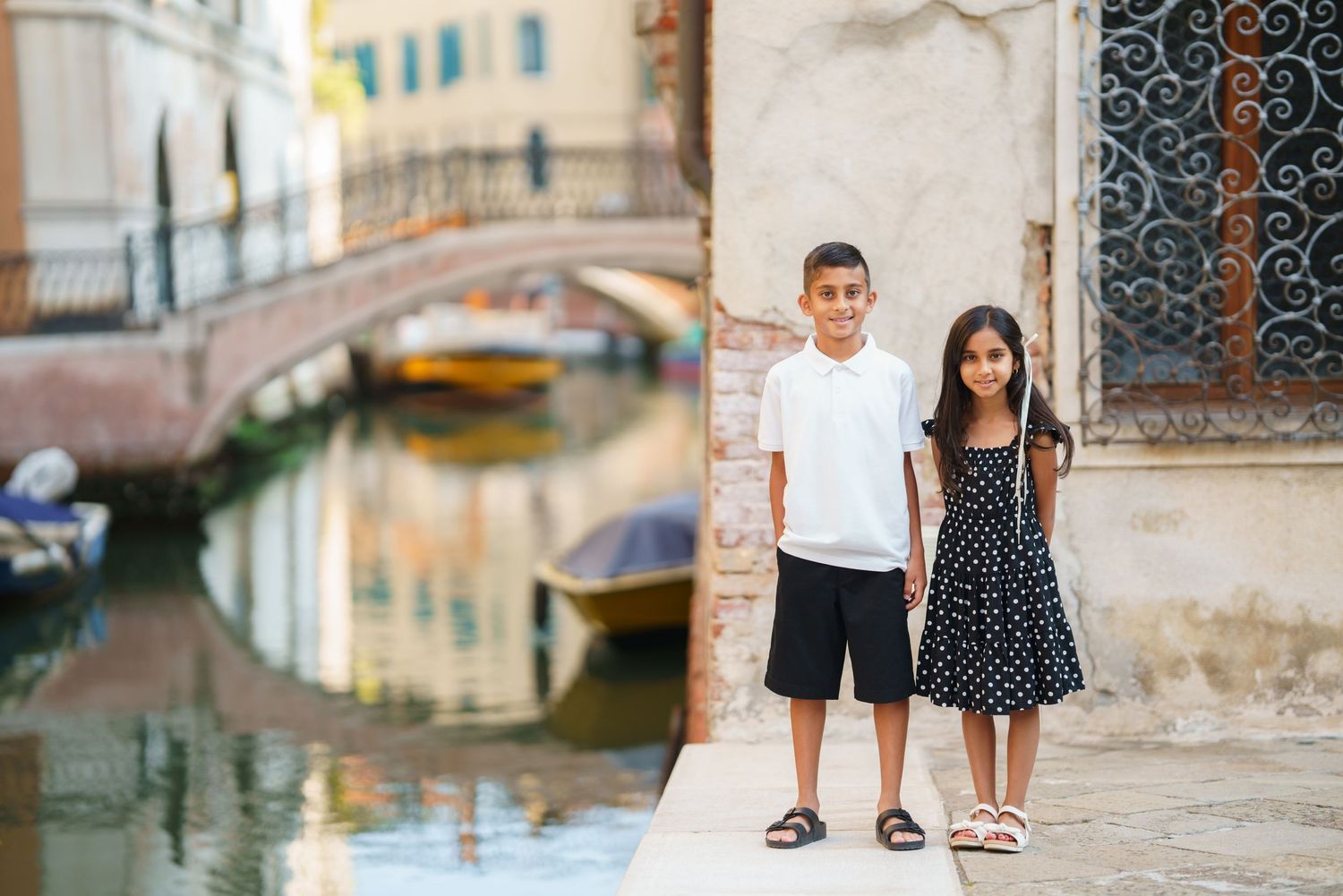 Two children pose on a Venice canal walkway with a historic bridge and moored boats in the background.