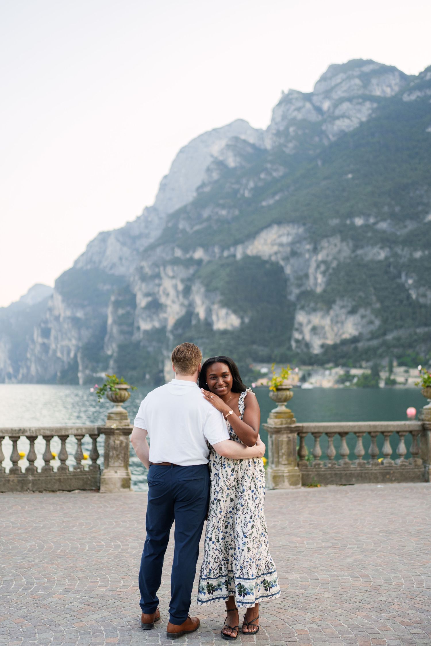 Woman smiling over her shoulder while embracing partner on a stone terrace with alpine lake views.