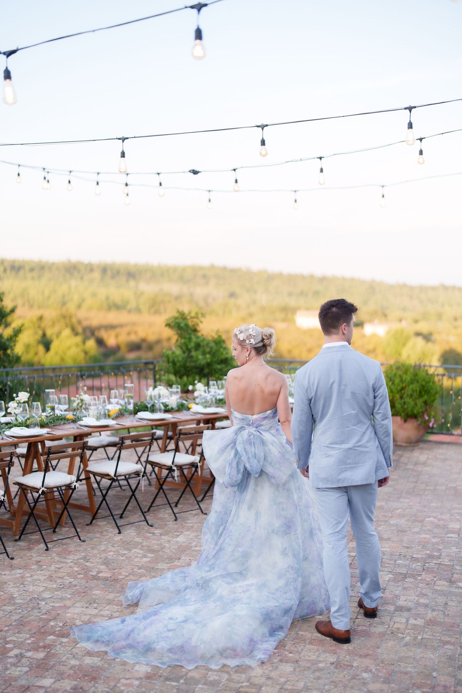 Bride in blue floral gown and groom in light suit walk toward outdoor wedding reception table under string lights.