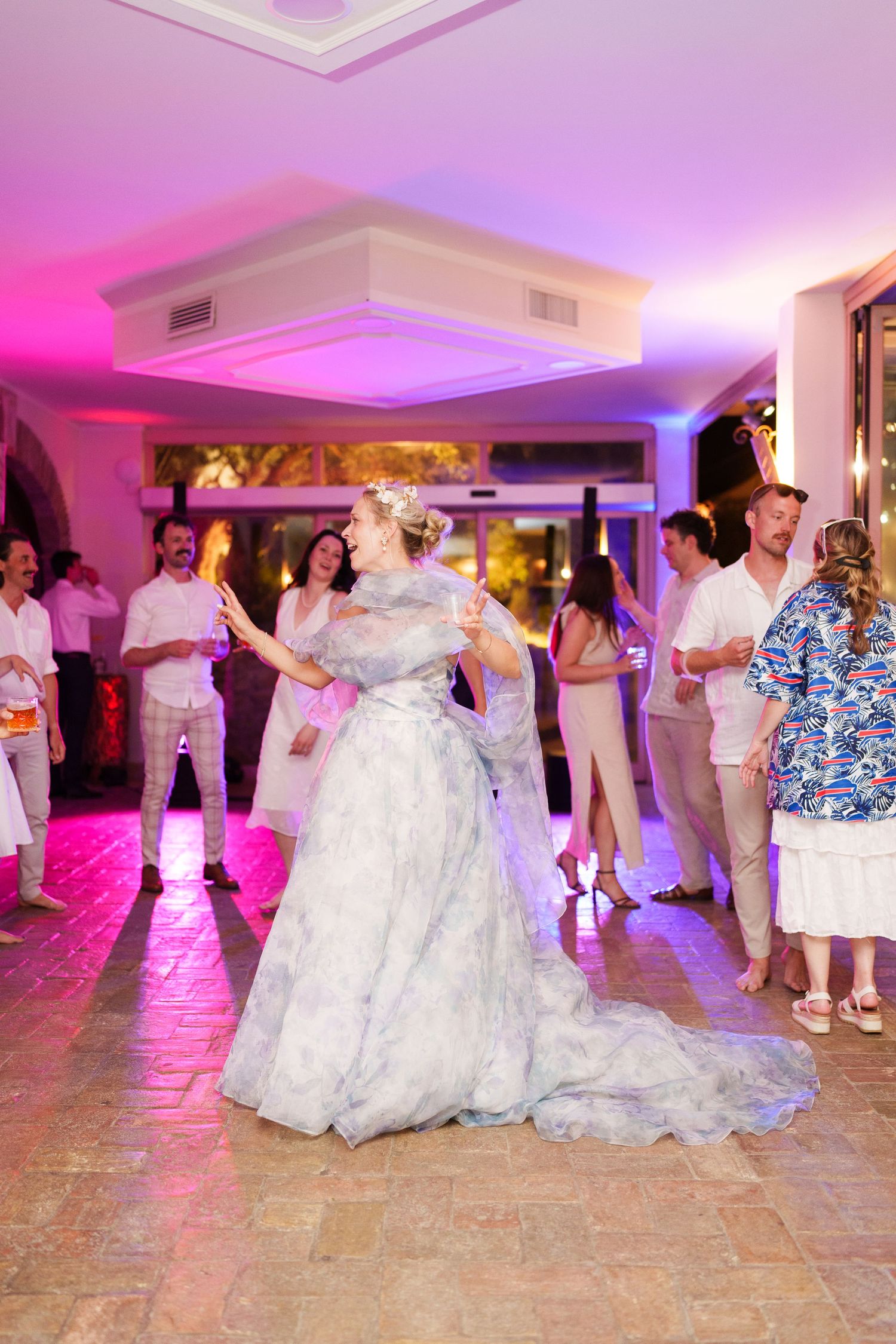 Bride in a blue floral gown dancing joyfully at her wedding reception with guests in the background.