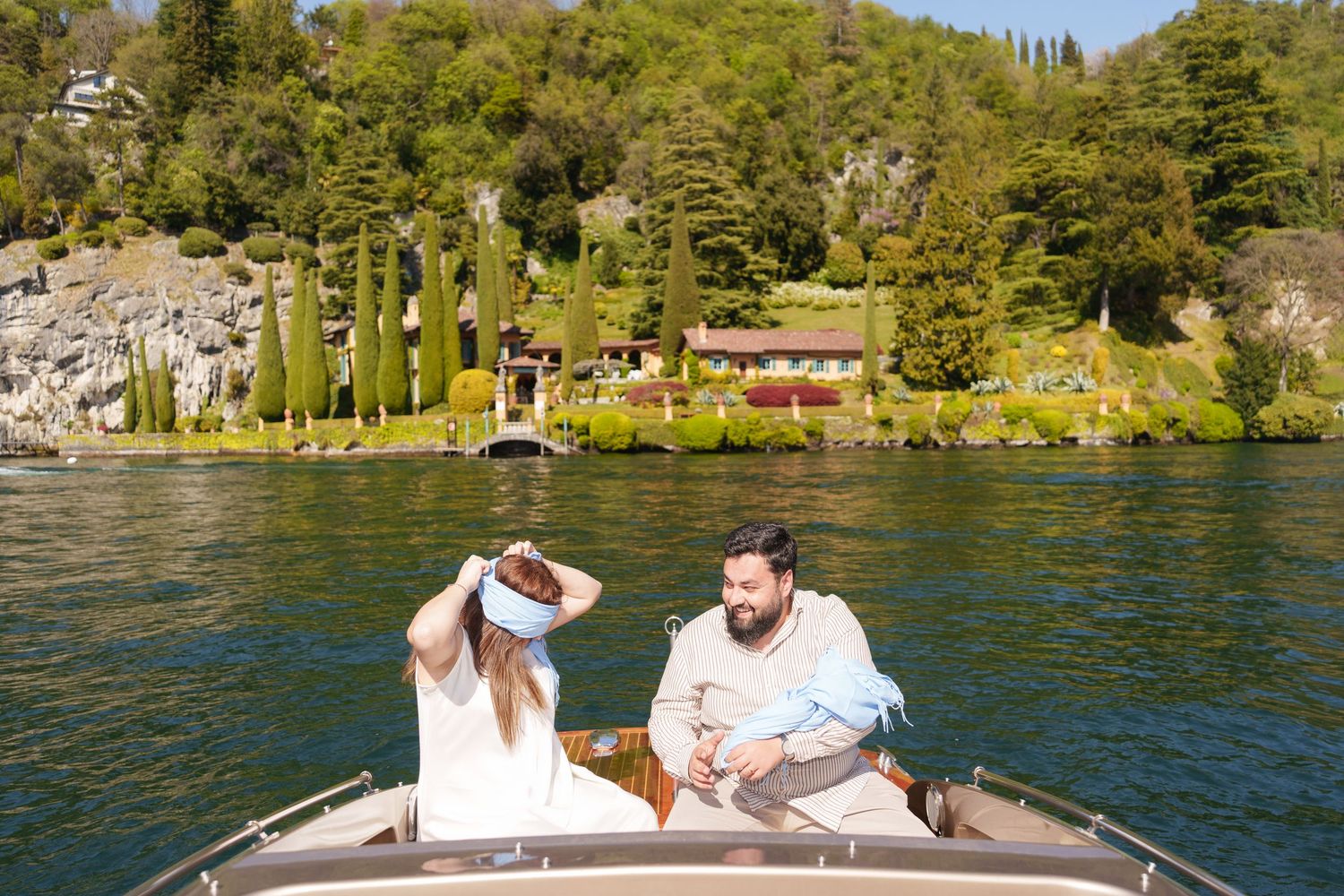 Couple relaxing on a boat on a scenic lake with lush green trees and a villa on the shore.