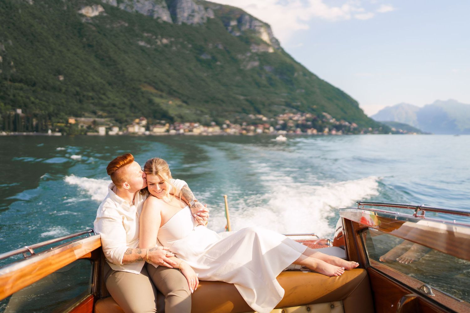 Couple in wedding attire relaxing on a wooden boat on Lake Como, Italy, with mountains and village in background.