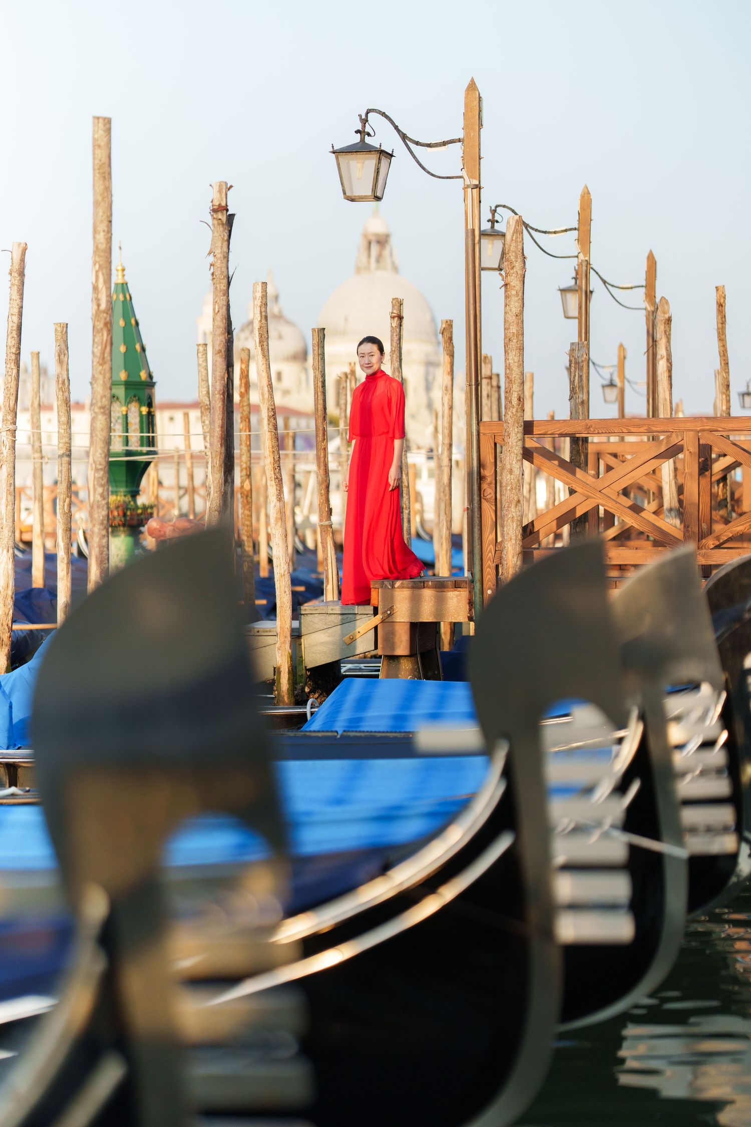 Woman in red dress standing on Venice gondola dock surrounded by moored gondolas with church dome in background.
