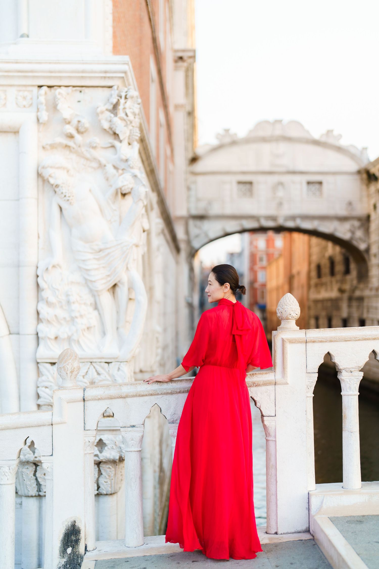 Woman in red gown stands on Venice balcony near Bridge of Sighs, Italy.