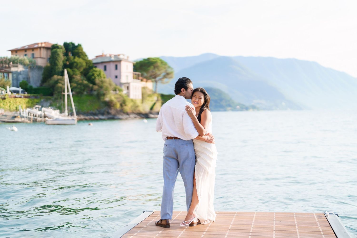Couple embraces on a dock overlooking serene lake with mountains and village in the background.