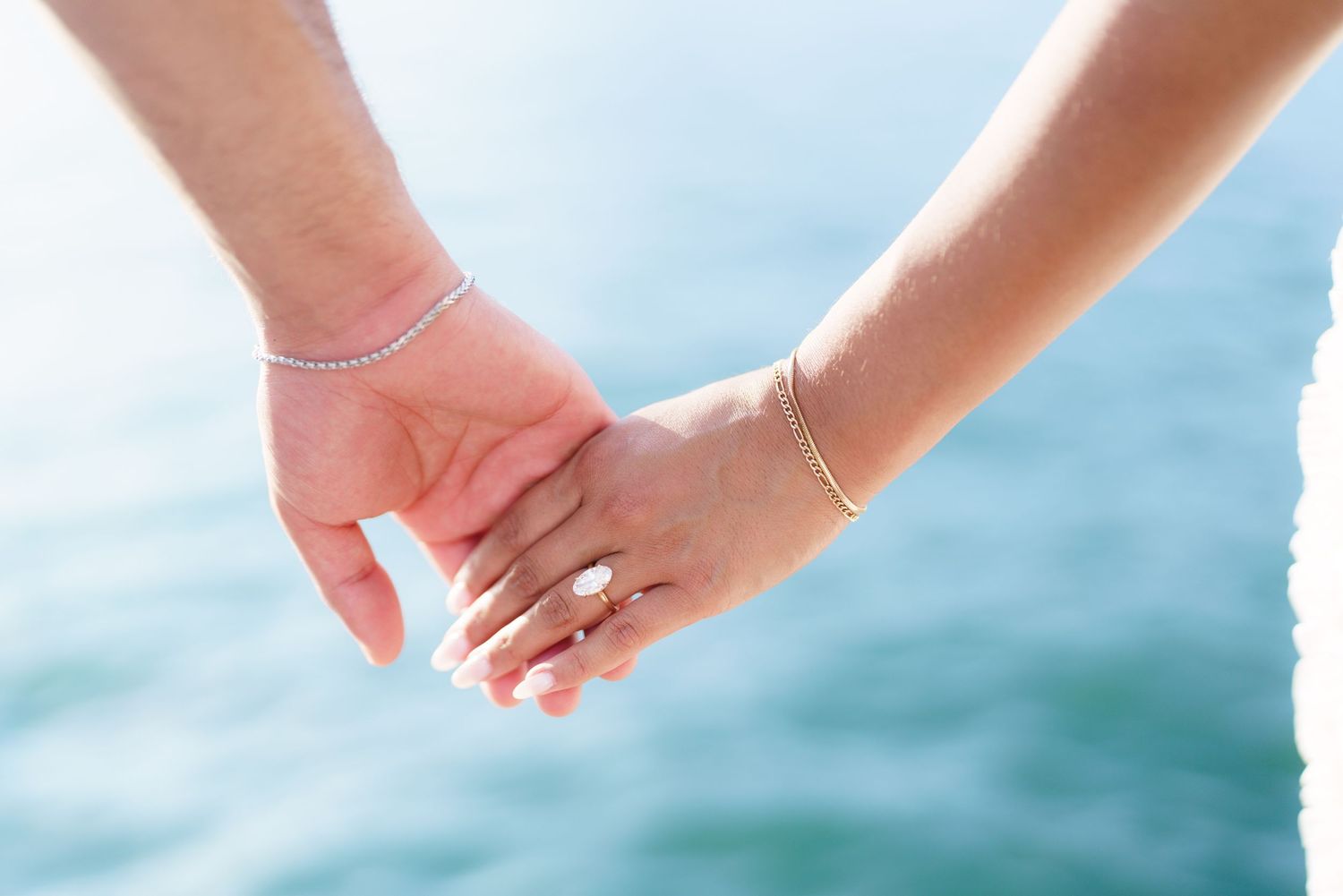 Couple holding hands near water, woman wearing diamond engagement ring and gold bracelet, romantic outdoor setting.