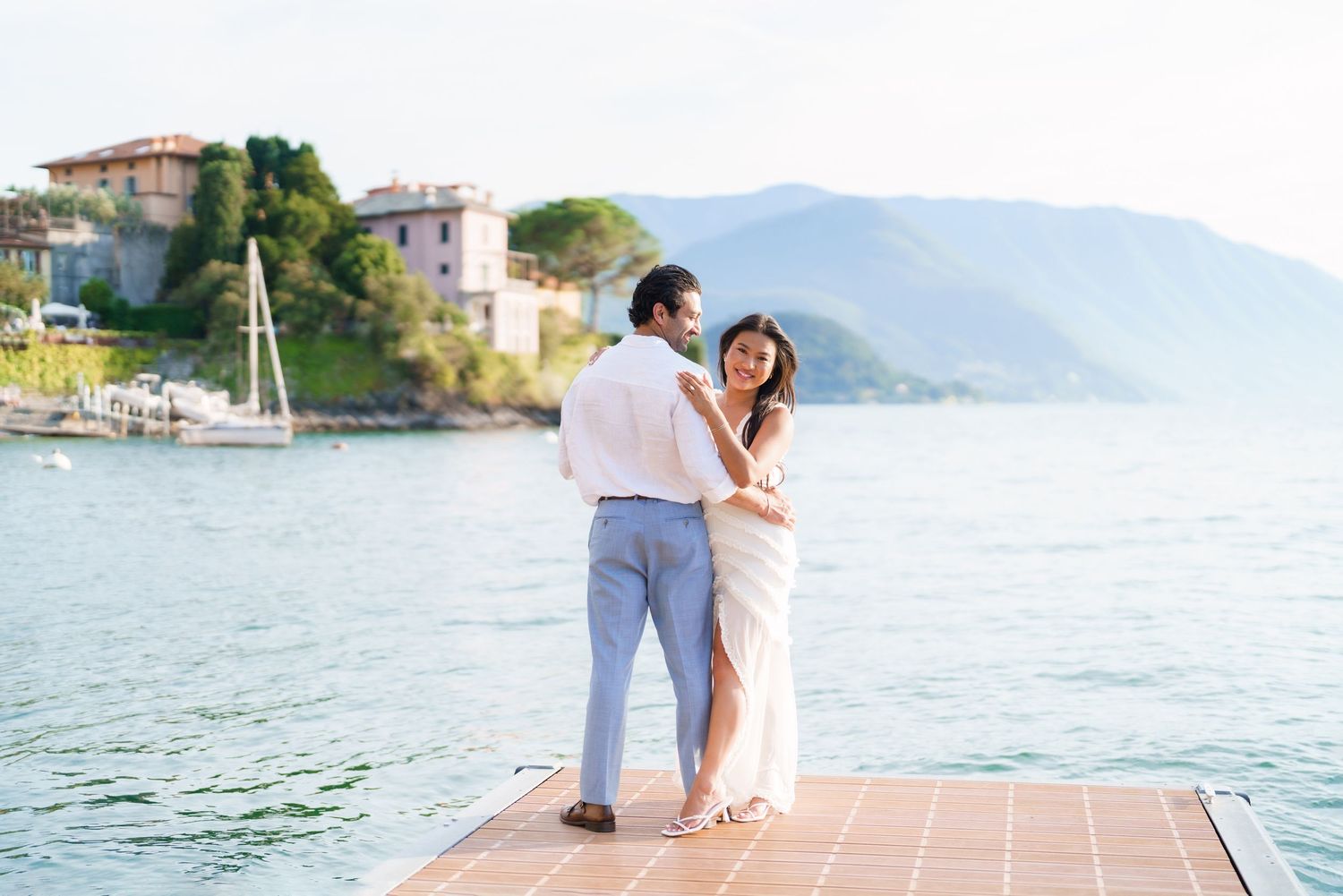 Couple in white outfits embracing on a dock overlooking a serene lake with mountains and a villa in Italy.