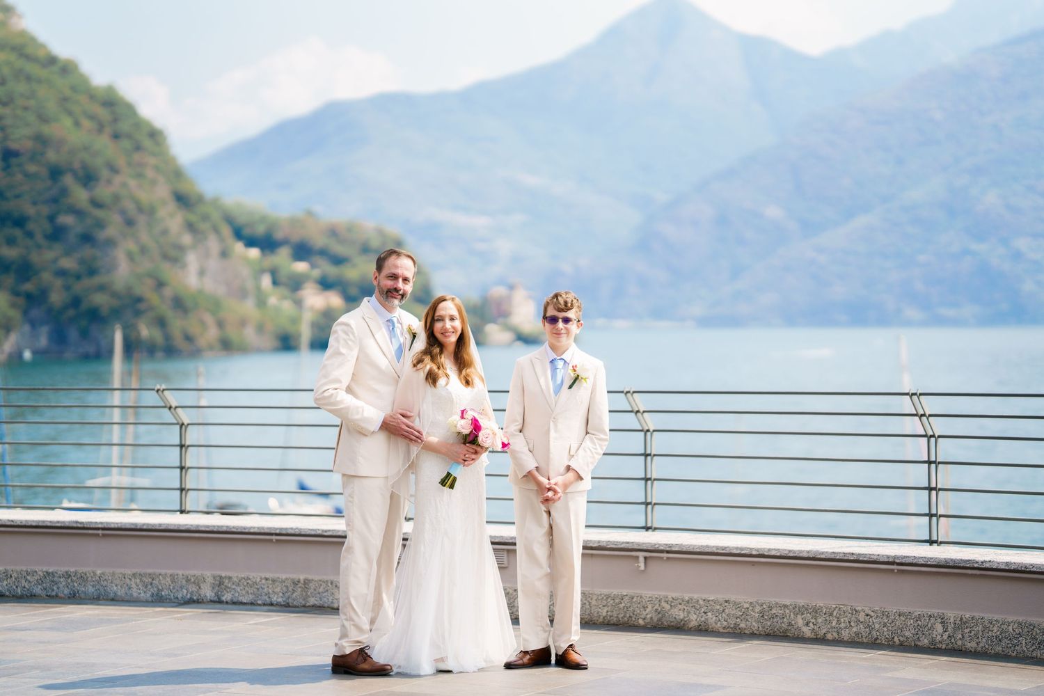 Bride and groom posing with guest in cream suits by a lakeside terrace with mountain views.