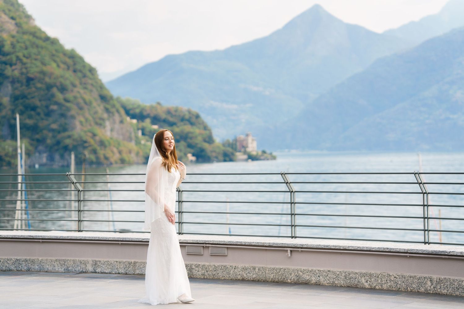 Bride in white gown poses on lakeside terrace with mountain backdrop at Lake Como, Italy.