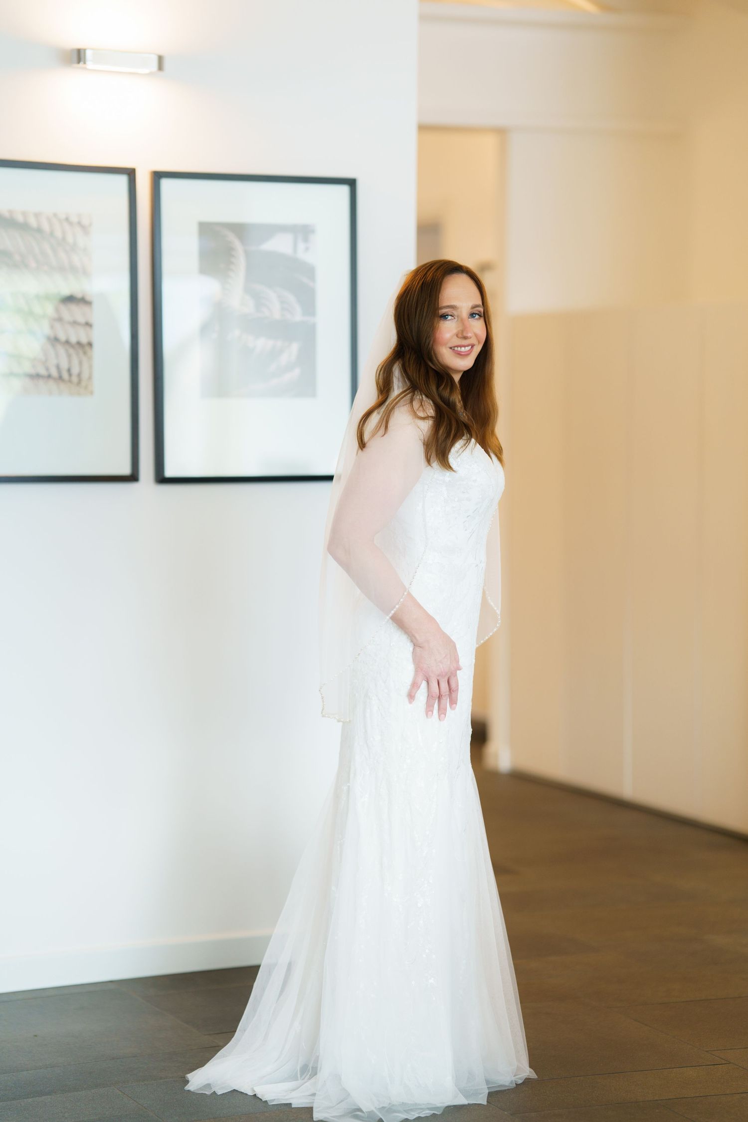 Bride in elegant white lace wedding gown with tulle skirt posing in a bright modern room with framed artwork.