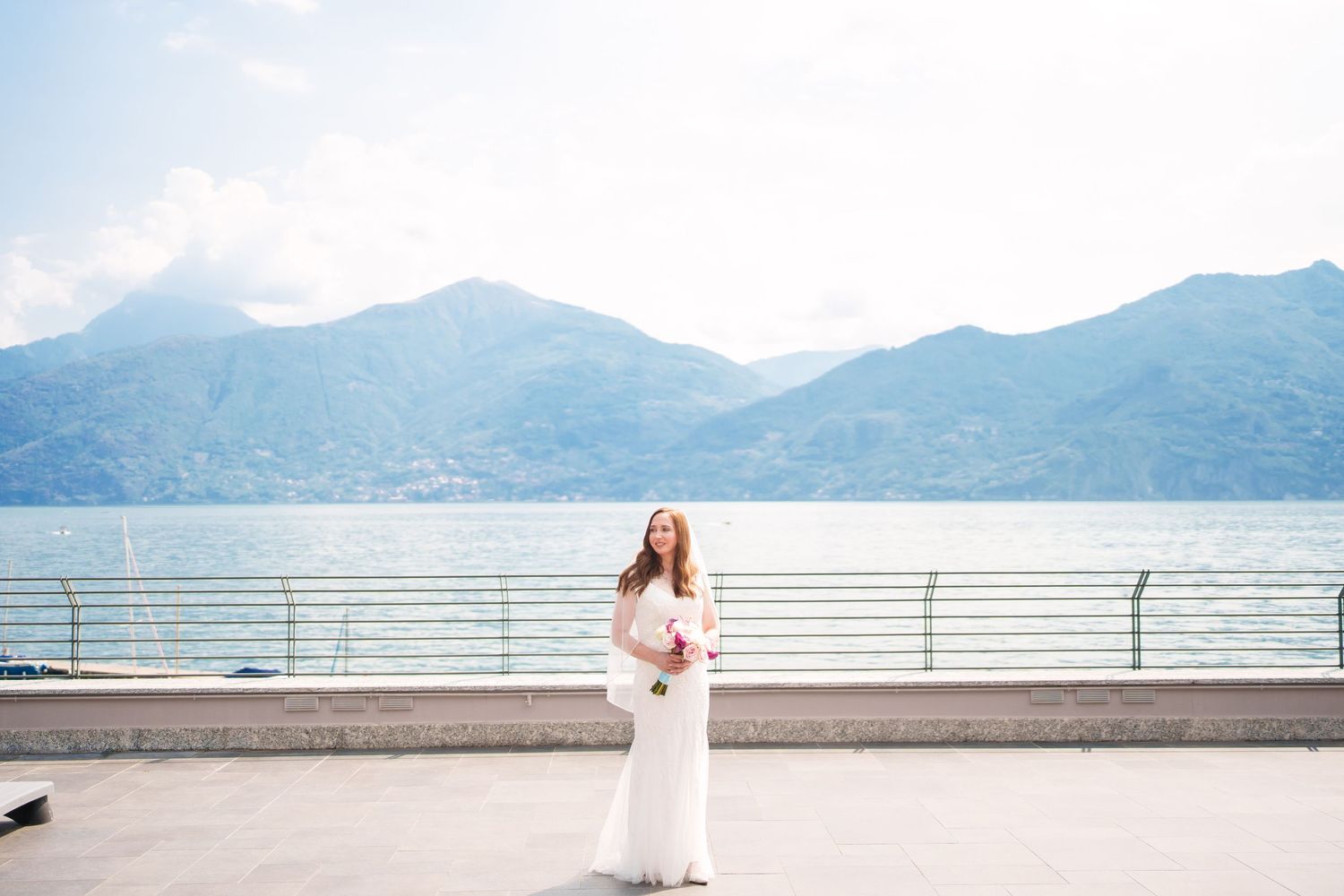Bride in white gown poses on lakeside promenade with mountain backdrop and serene blue waters.