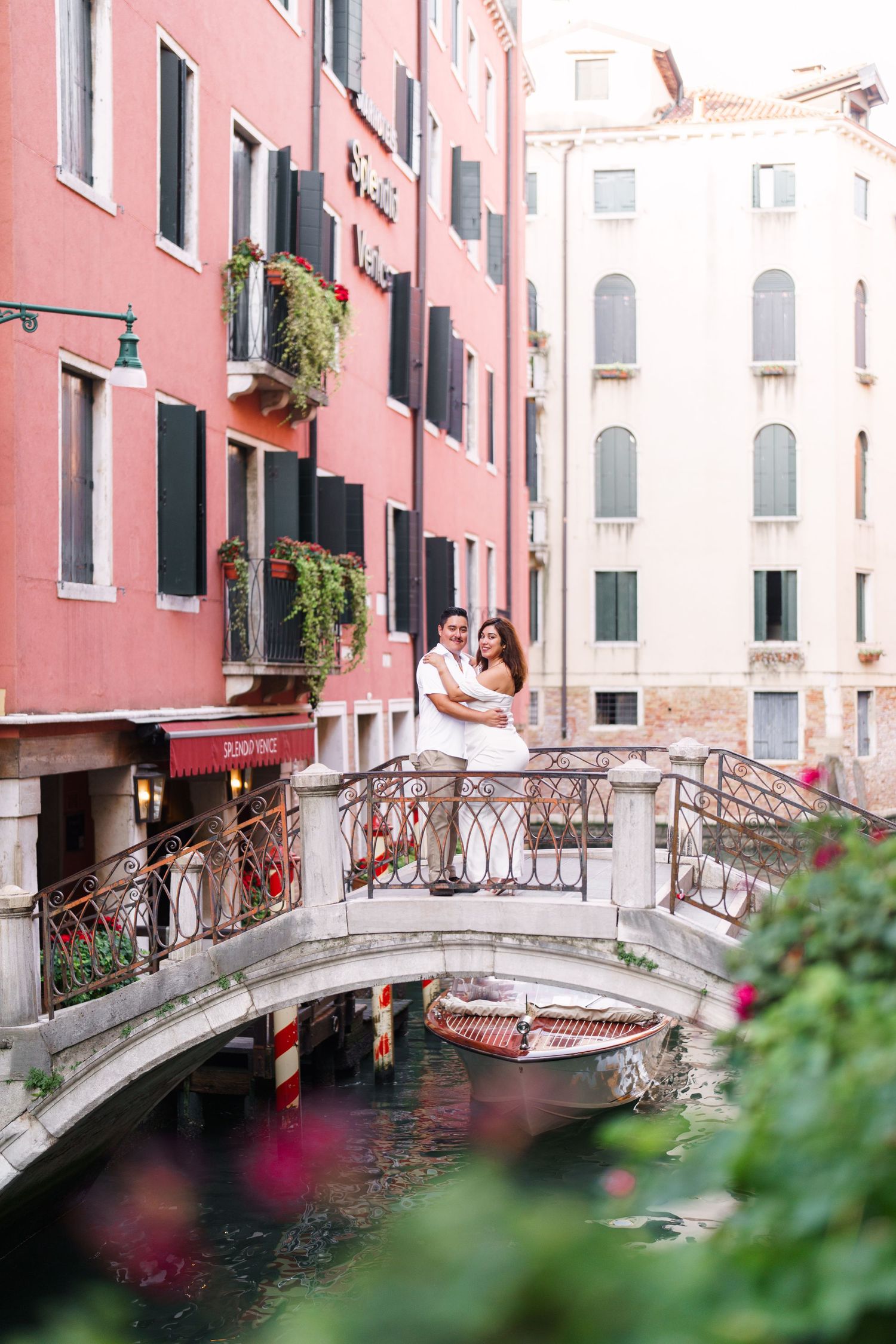 A romantic couple stands on a charming footbridge over a narrow canal in Venice with pink buildings in the background.