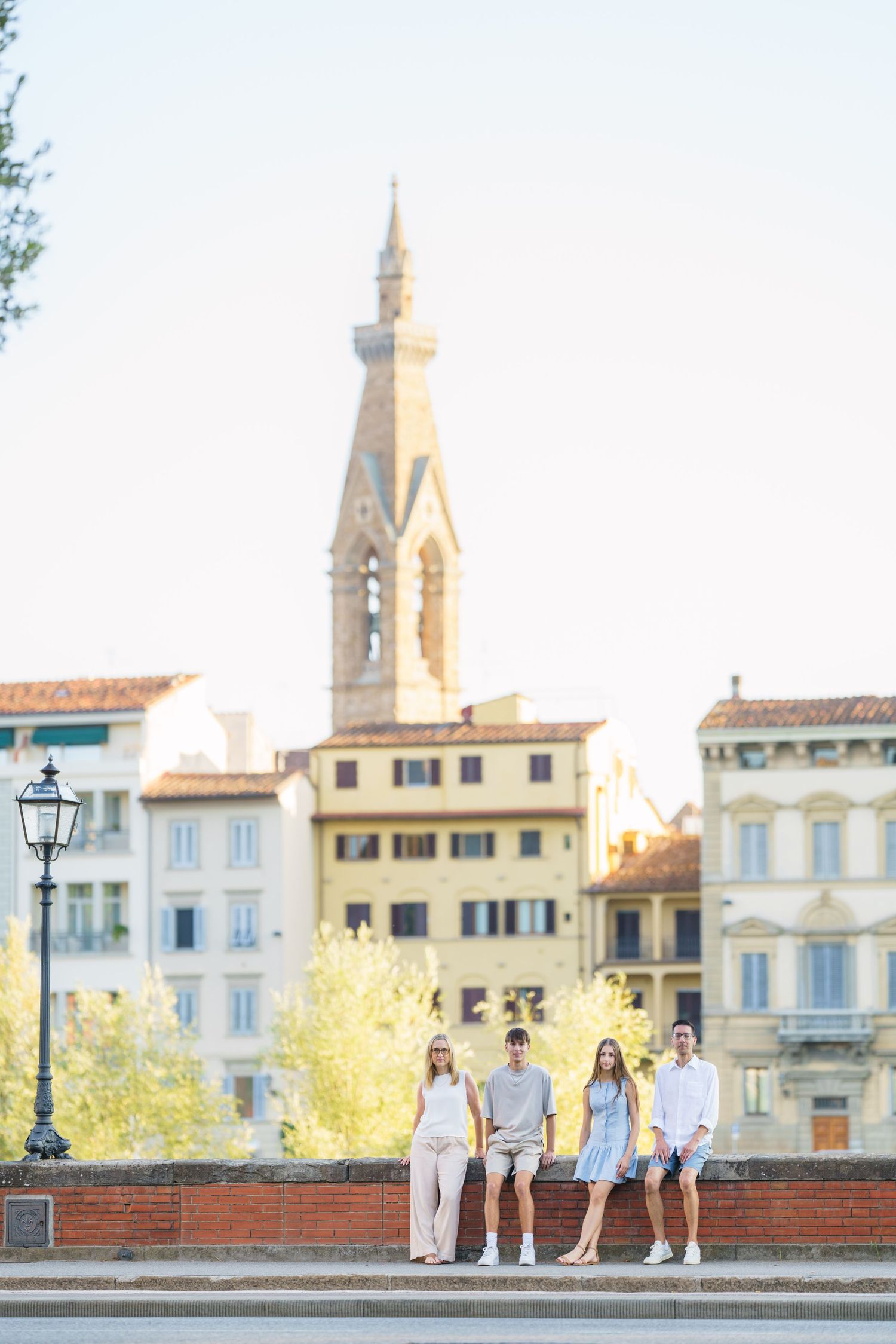A group of people walking along a historic stone wall with classic Italian architecture and church spires in the background.