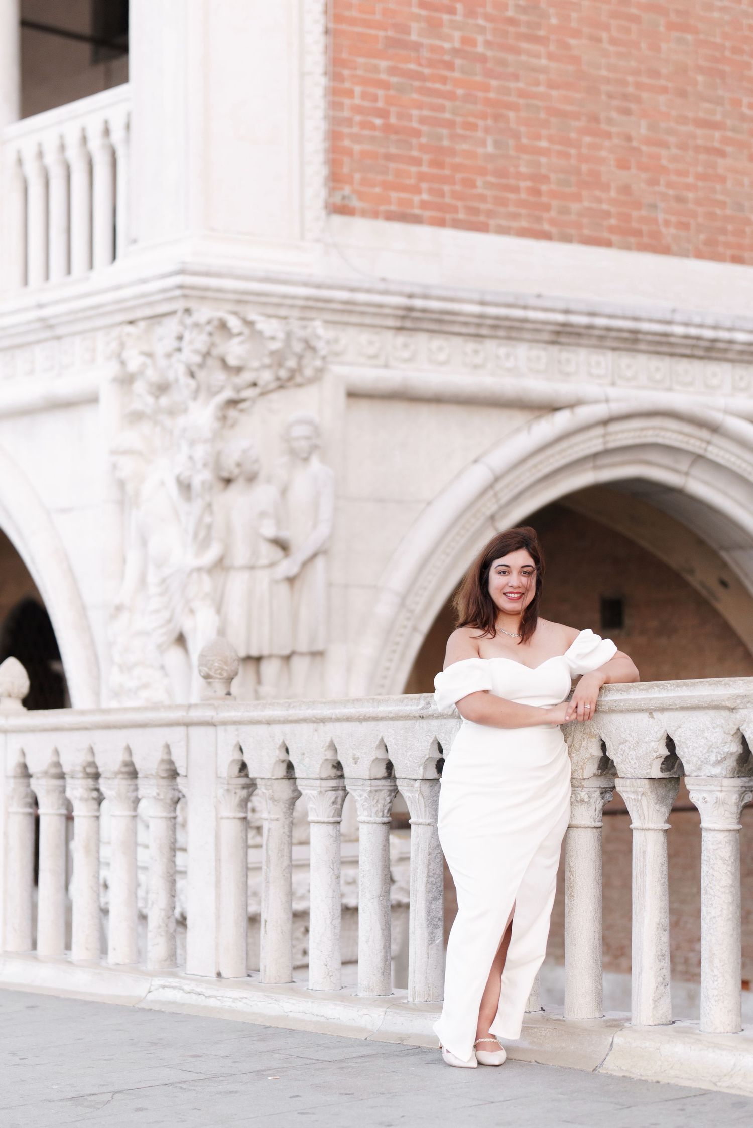 A bride in a white dress poses along an ornate white stone balcony against a brick wall background.