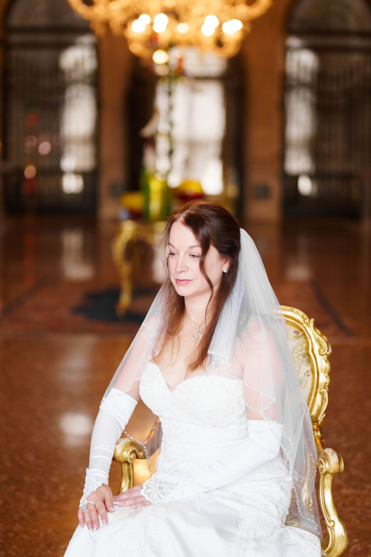 A bride in a white wedding dress sits elegantly on an ornate gold chair in a grand ballroom setting.