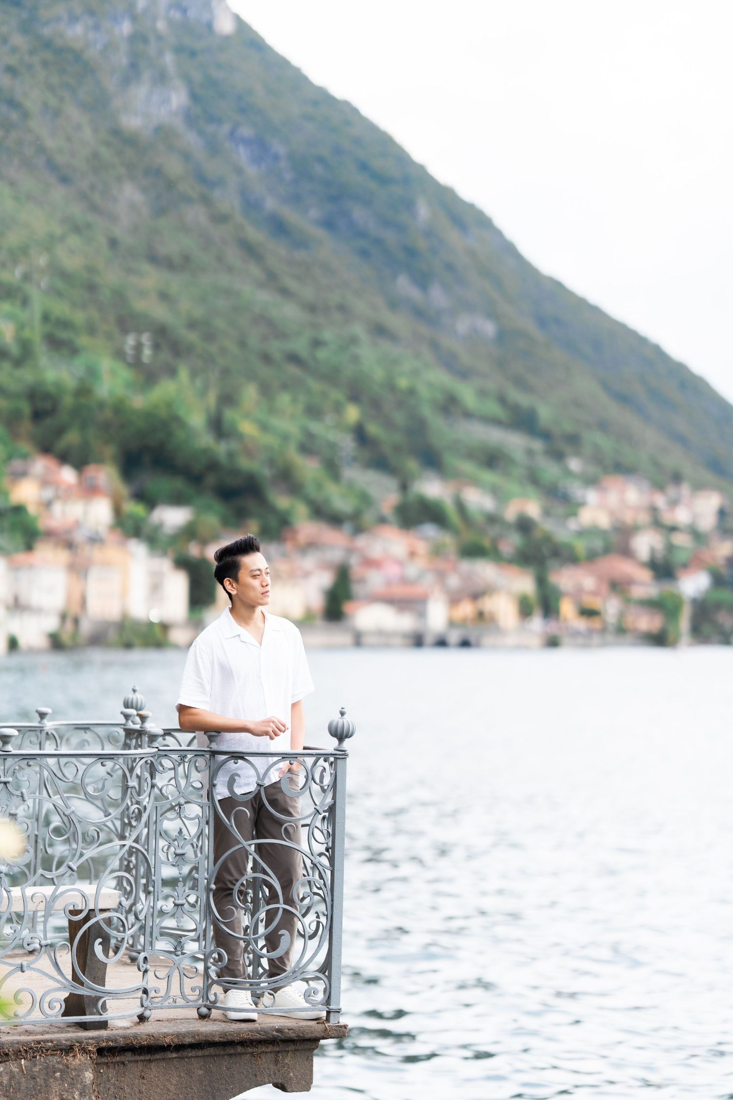 A person sits on an ornate metal bench overlooking Lake Como with mountains and Italian village buildings in background.
