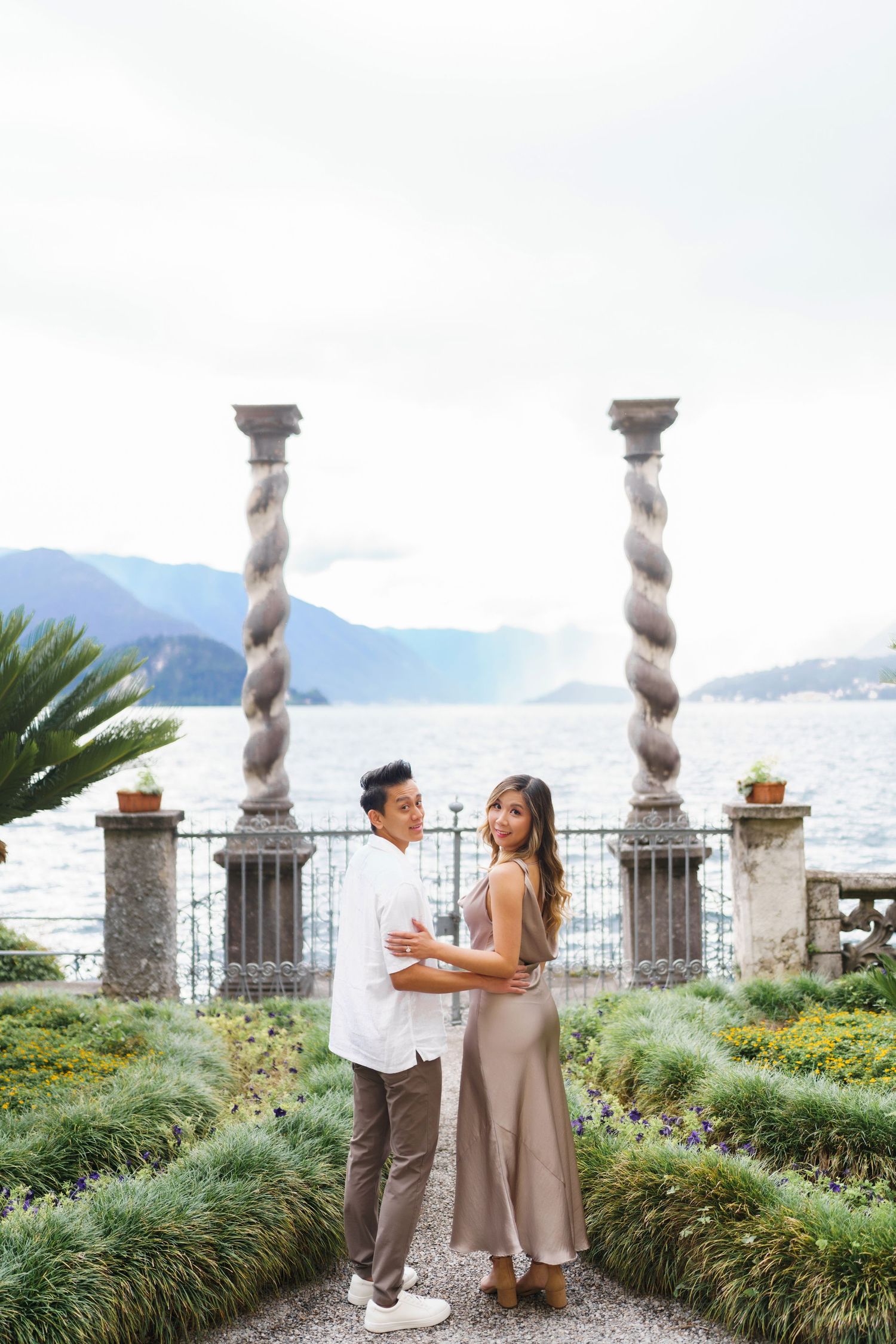 A romantic couple shares an intimate moment on a scenic terrace overlooking Lake Como with ornate stone columns and gardens.