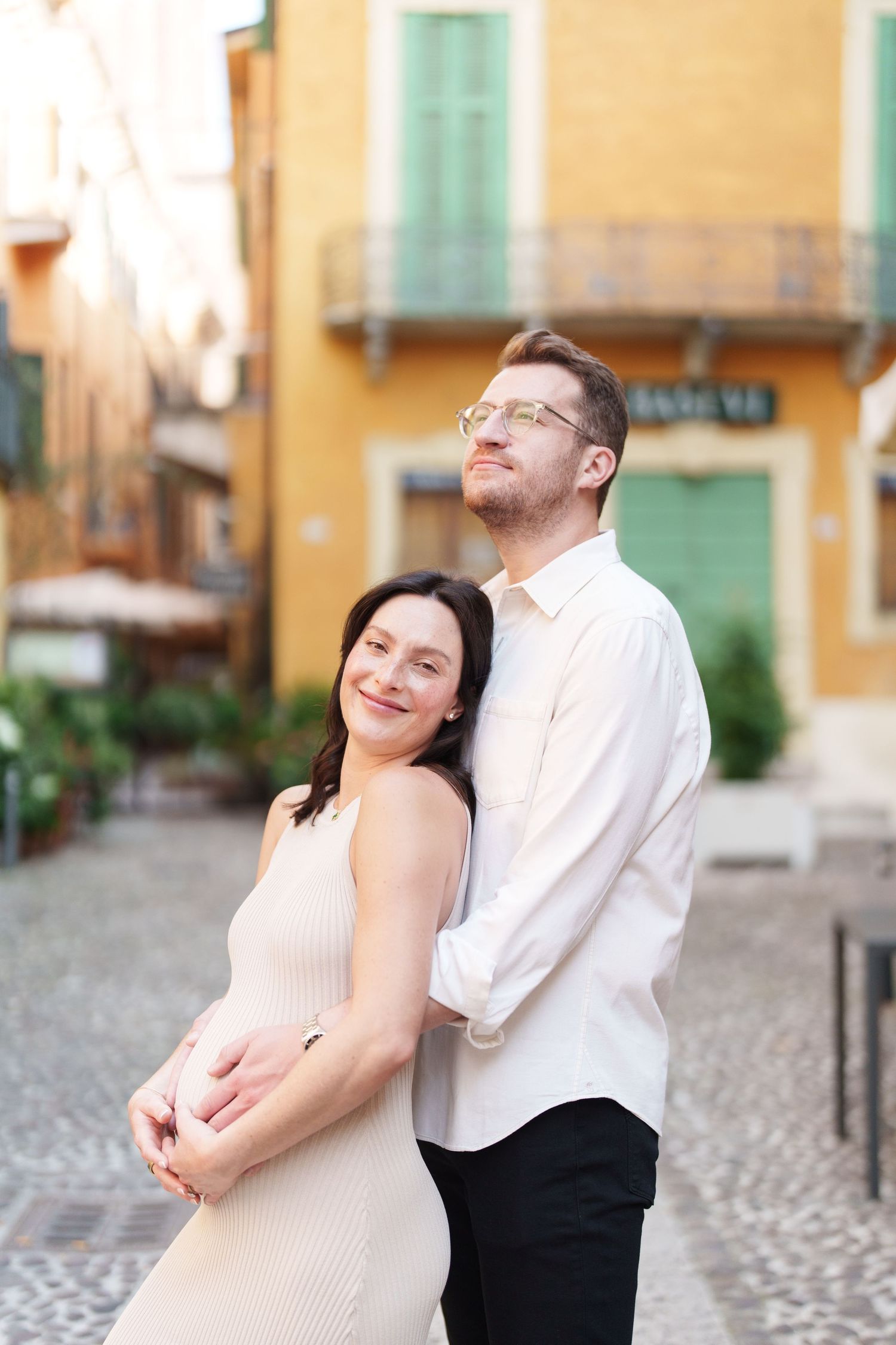 Couple shares tender moment while posing together on cobblestone street with Mediterranean architecture behind them.