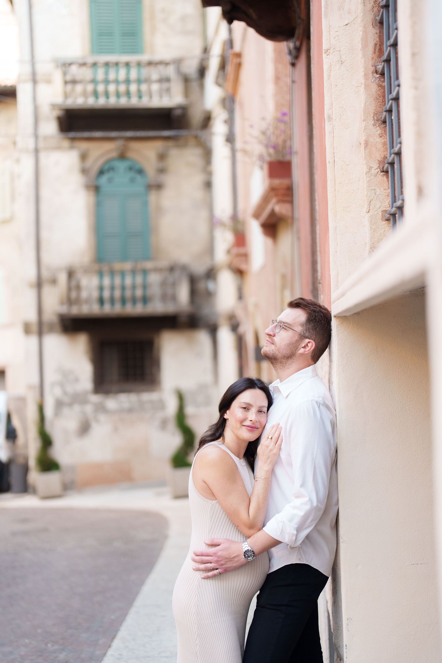 A couple shares an intimate moment together on a European street with turquoise shutters and stone walls in the background.