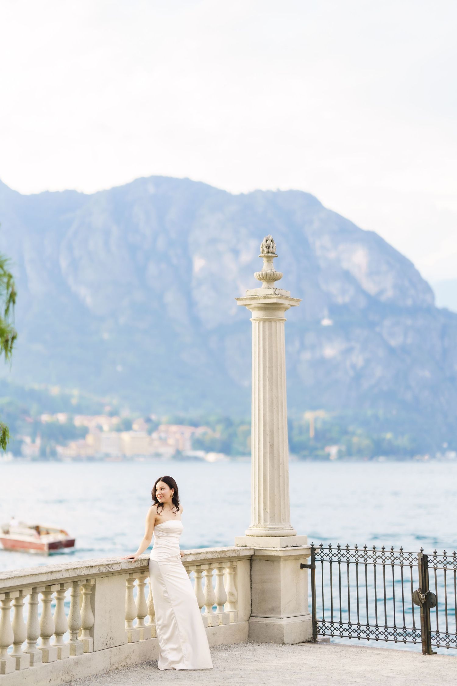 Elegant figure in white dress poses on lakeside terrace with ornate column and mountain backdrop at Lake Como, Italy.