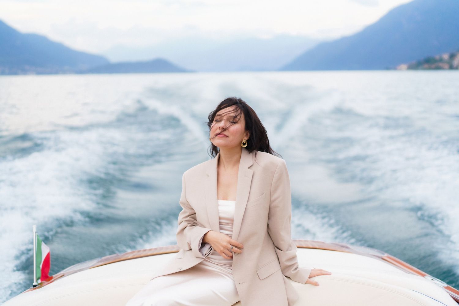A person in a beige blazer enjoys a boat ride on turquoise waters with mountains in the background.