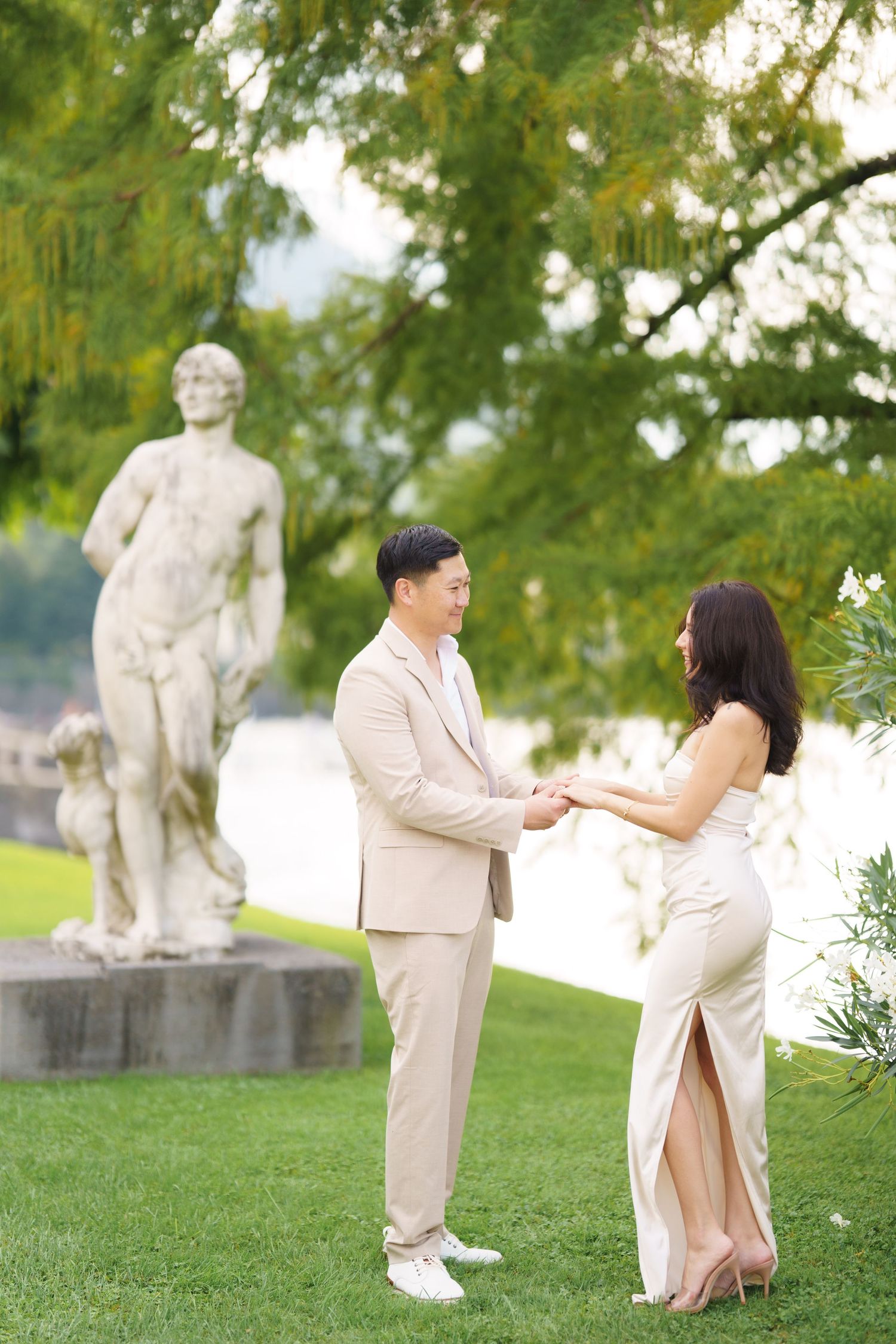 A couple in light colored outfits shares a romantic moment together in a park setting with green trees and a statue in background.