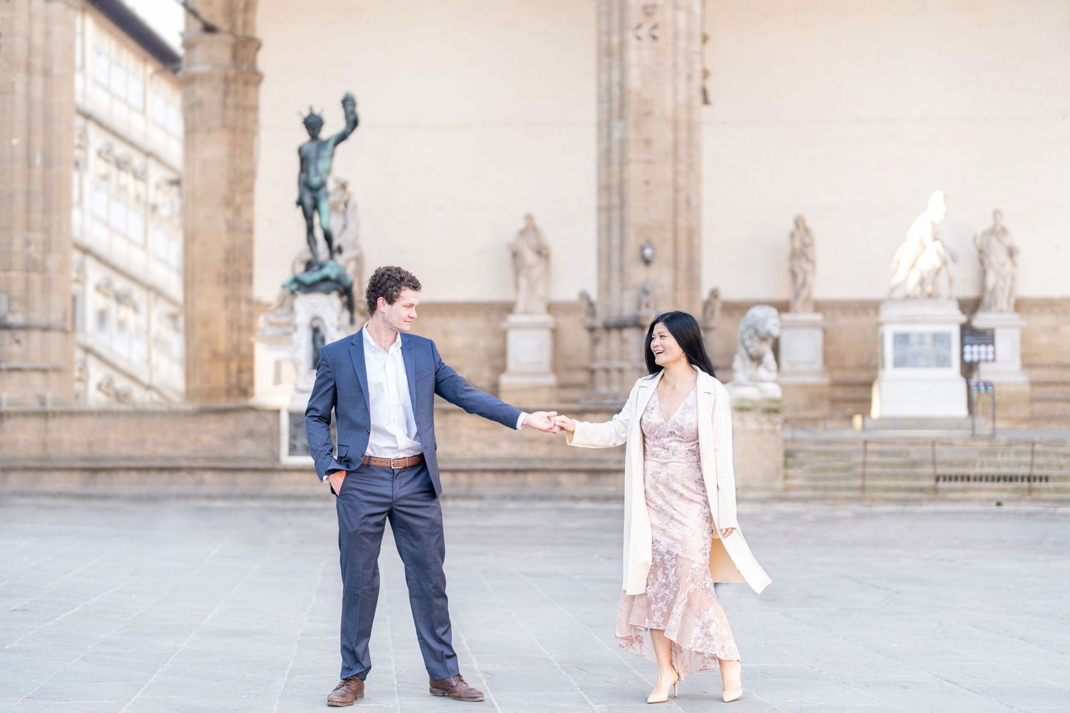 A romantic couple in formal attire dances and twirls together in front of a grand stone building with arches.