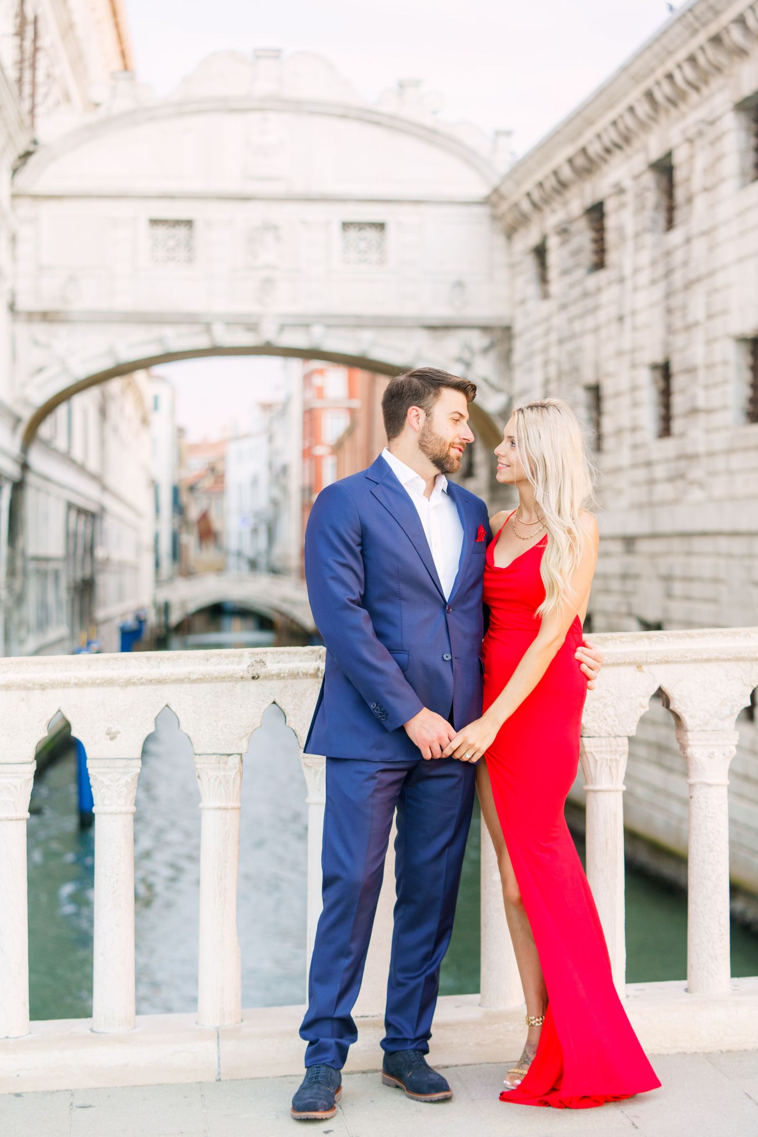 Romantic couple in red dress and navy suit pose along Venice's iconic Bridge of Sighs canal during photoshoot.
