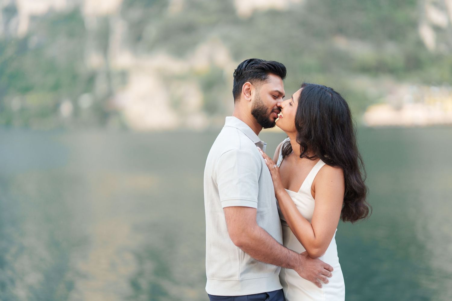A romantic couple embraces by a serene lakeside with mountains in the background during a photo session.