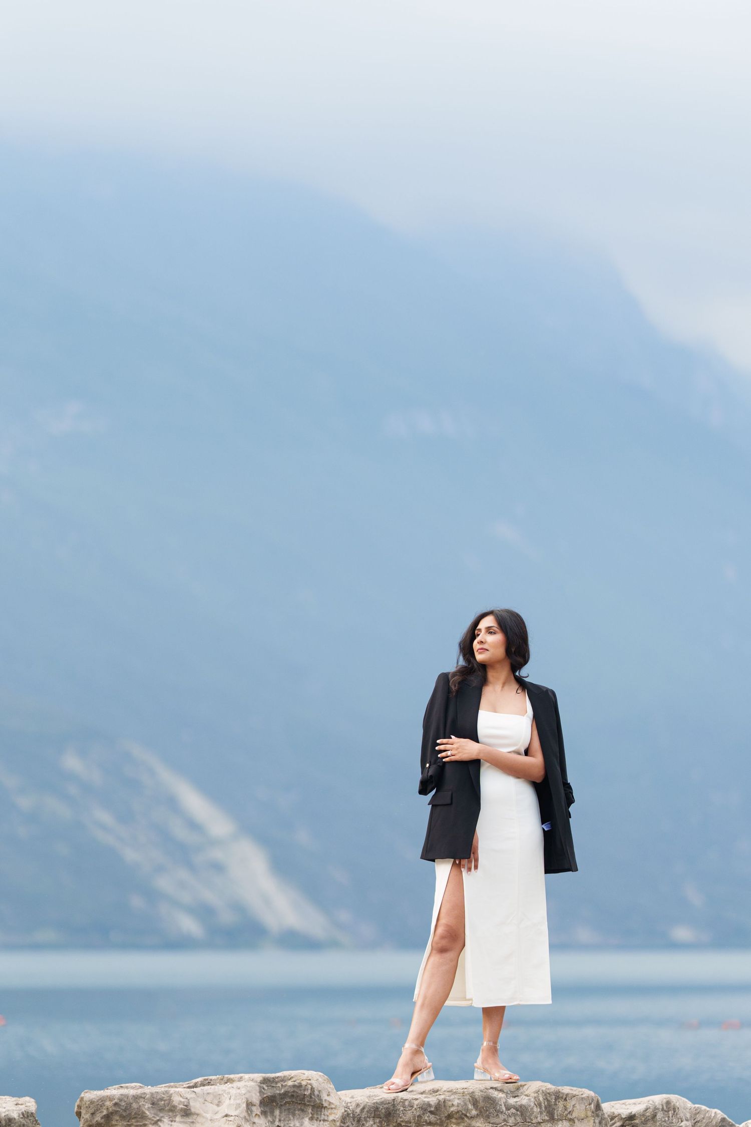 Couple in wedding attire embracing while standing on rocky coastal cliff overlooking ocean and mountains.