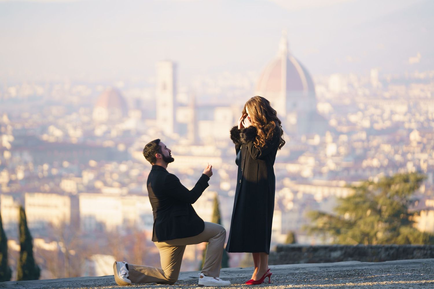 Two people sit together on a hilltop overlooking a scenic cityscape during sunset.