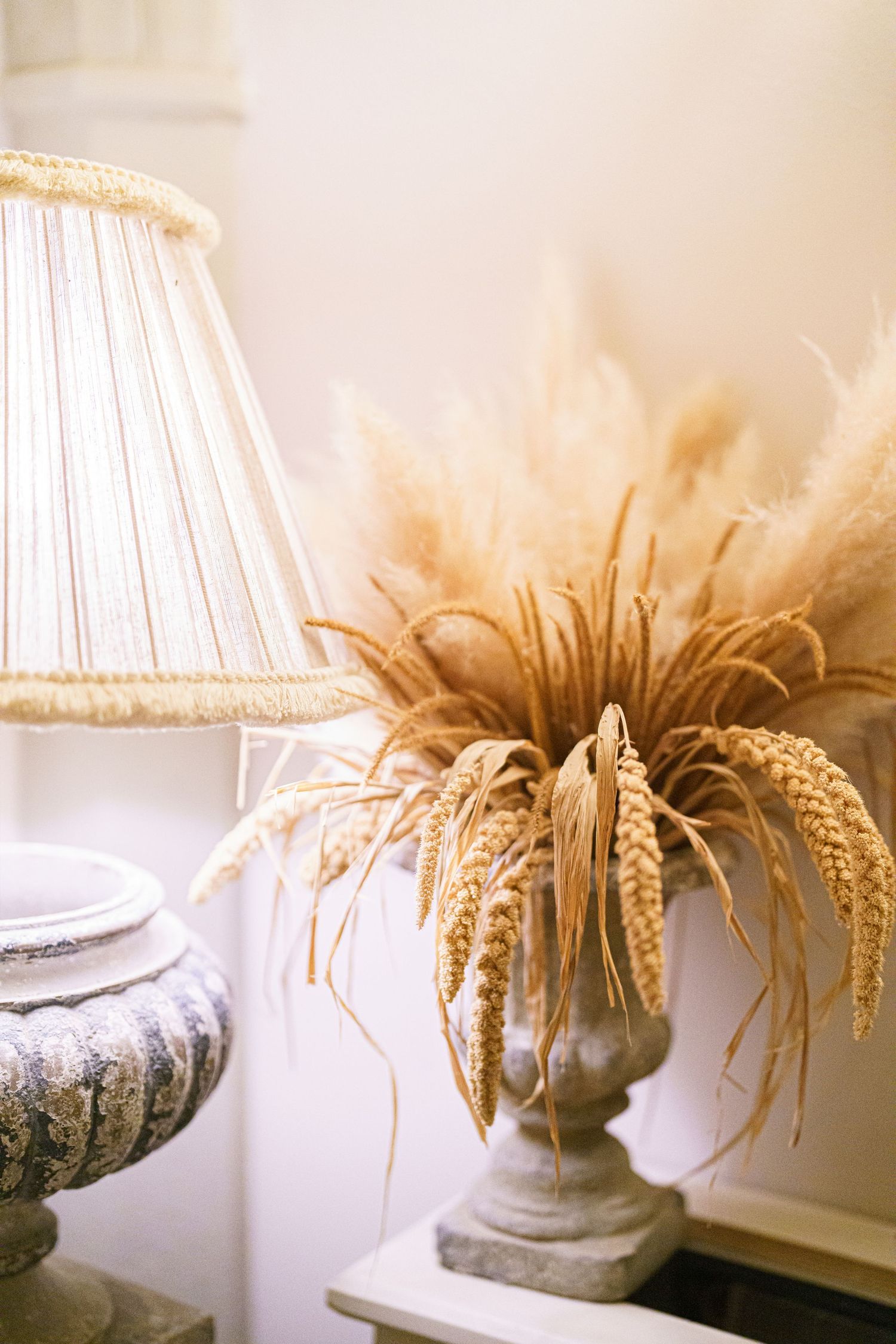 Elegant table lamp with pleated shade next to dried wheat arrangement in stone vase against neutral wall.