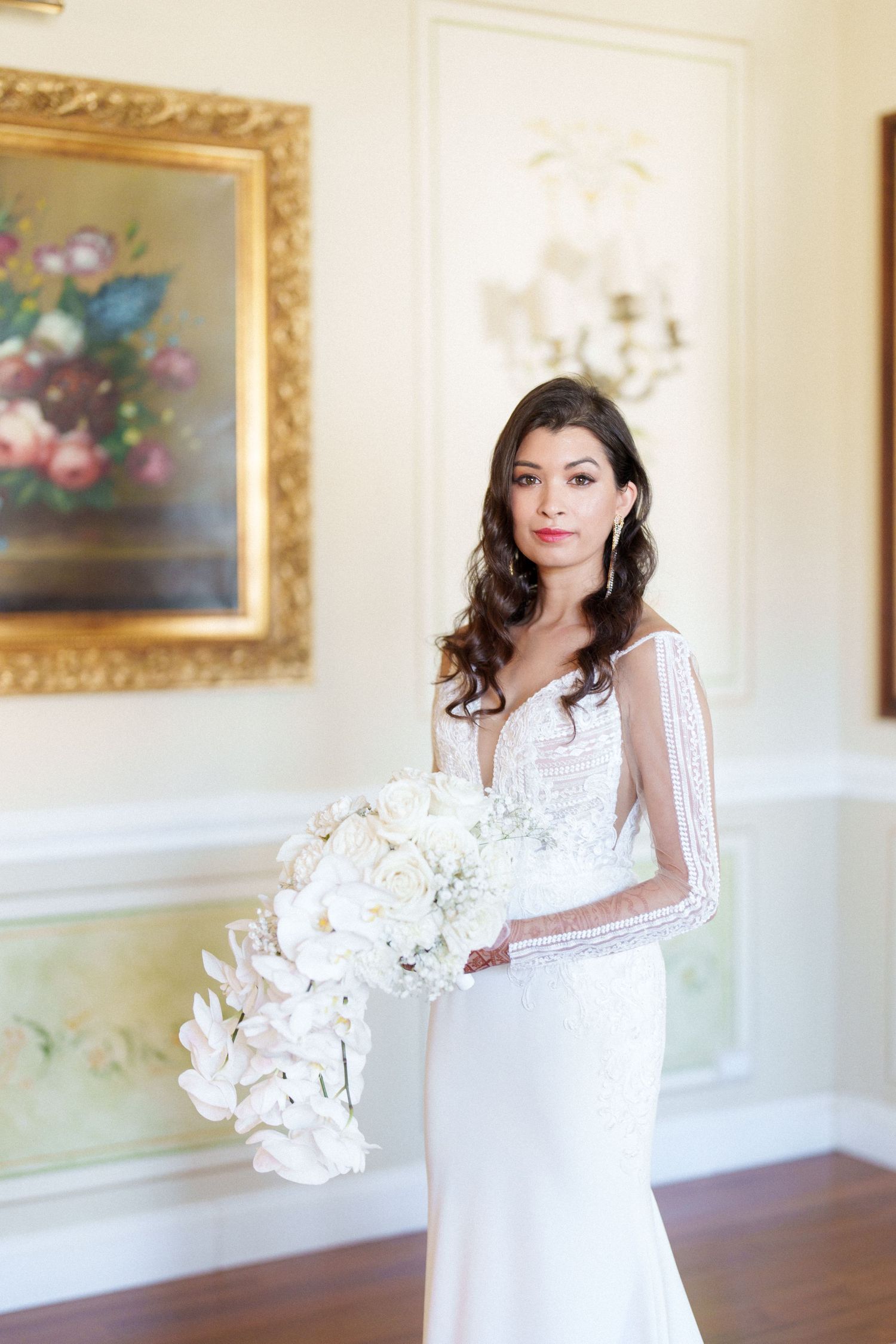 Bride in elegant white gown holds bouquet in room with ornate gold-framed artwork.