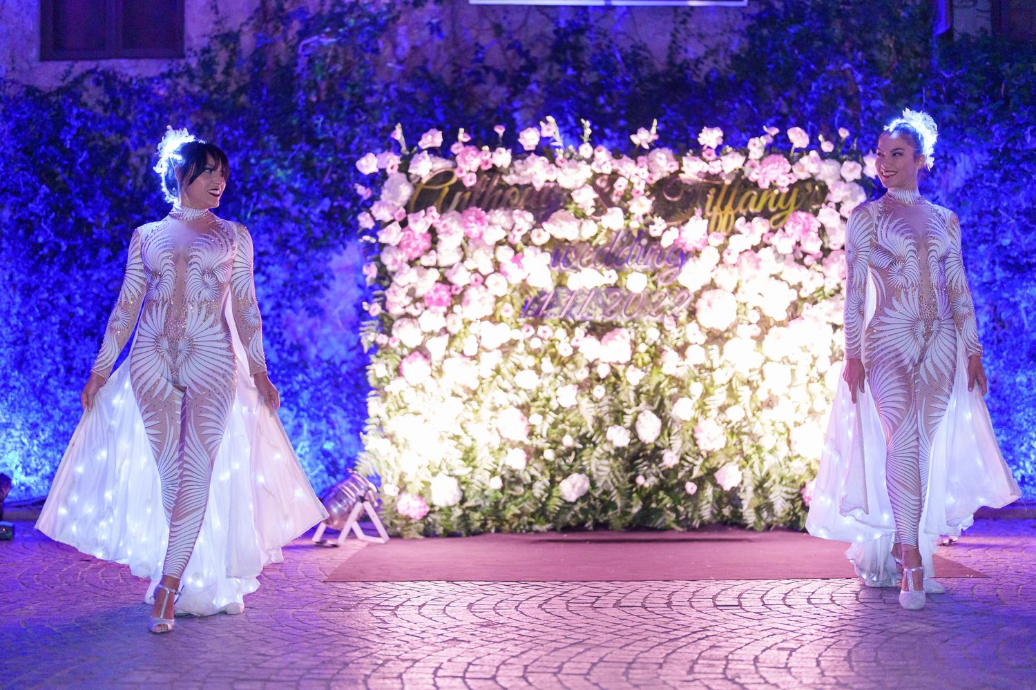 Elegant wedding party members in white dresses walk gracefully under blue-lit floral decorations.