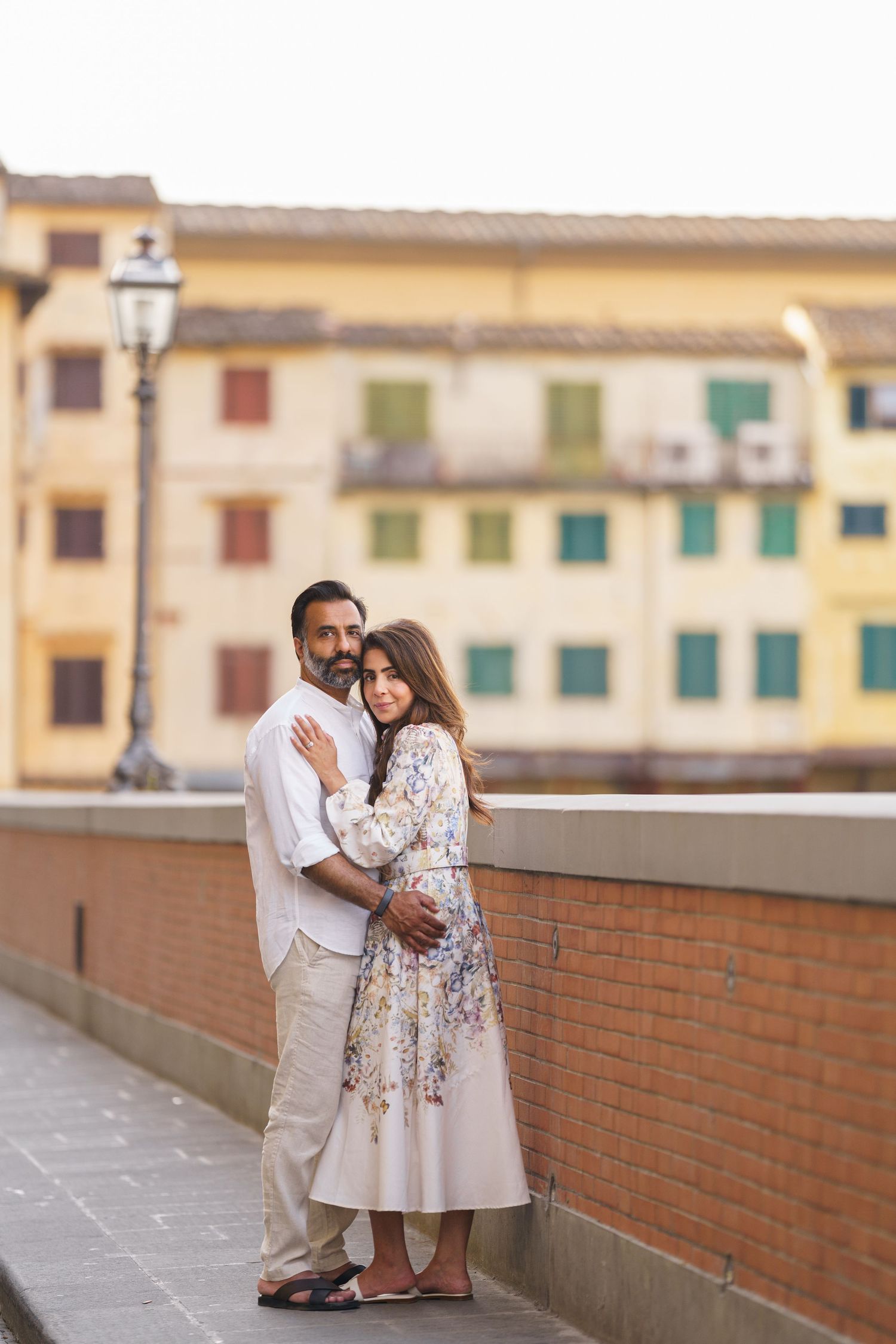 Couple embracing on a bridge in Pisa, Italy, with colorful buildings in the background.