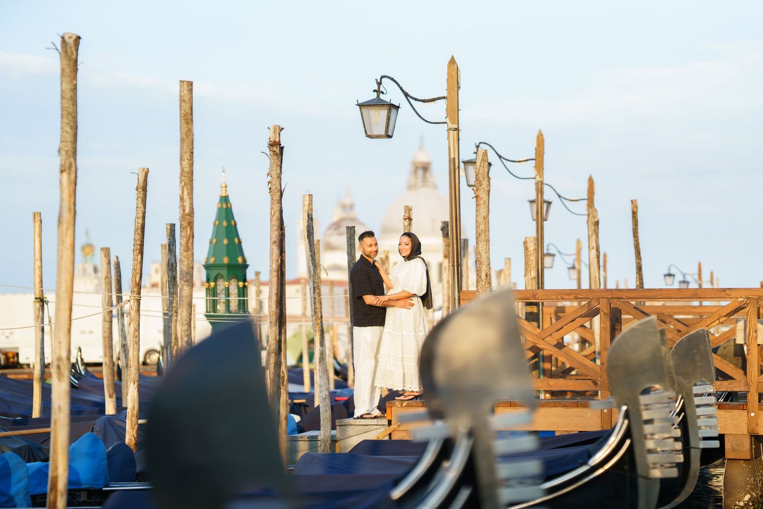 Couple embracing among gondolas and wooden poles at Venice's waterfront with historic architecture in background.
