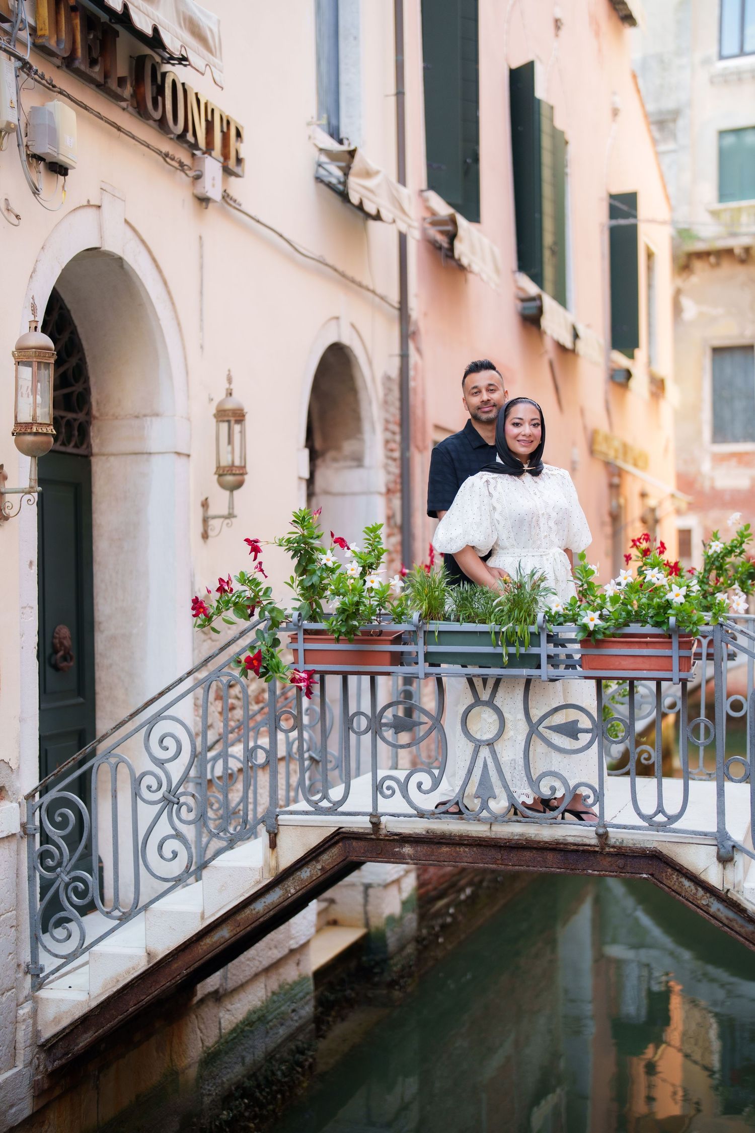 Couple standing on ornate bridge over Venice canal with colorful flowers and historic buildings backdrop.