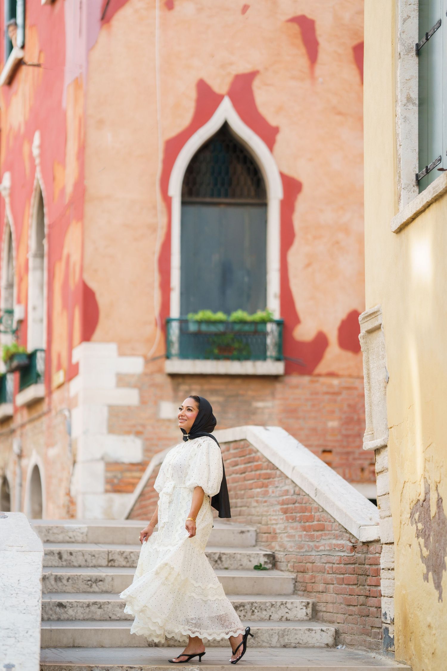 Woman in white lace dress ascending stone steps in Venice, Italy, with colorful historic buildings in background.