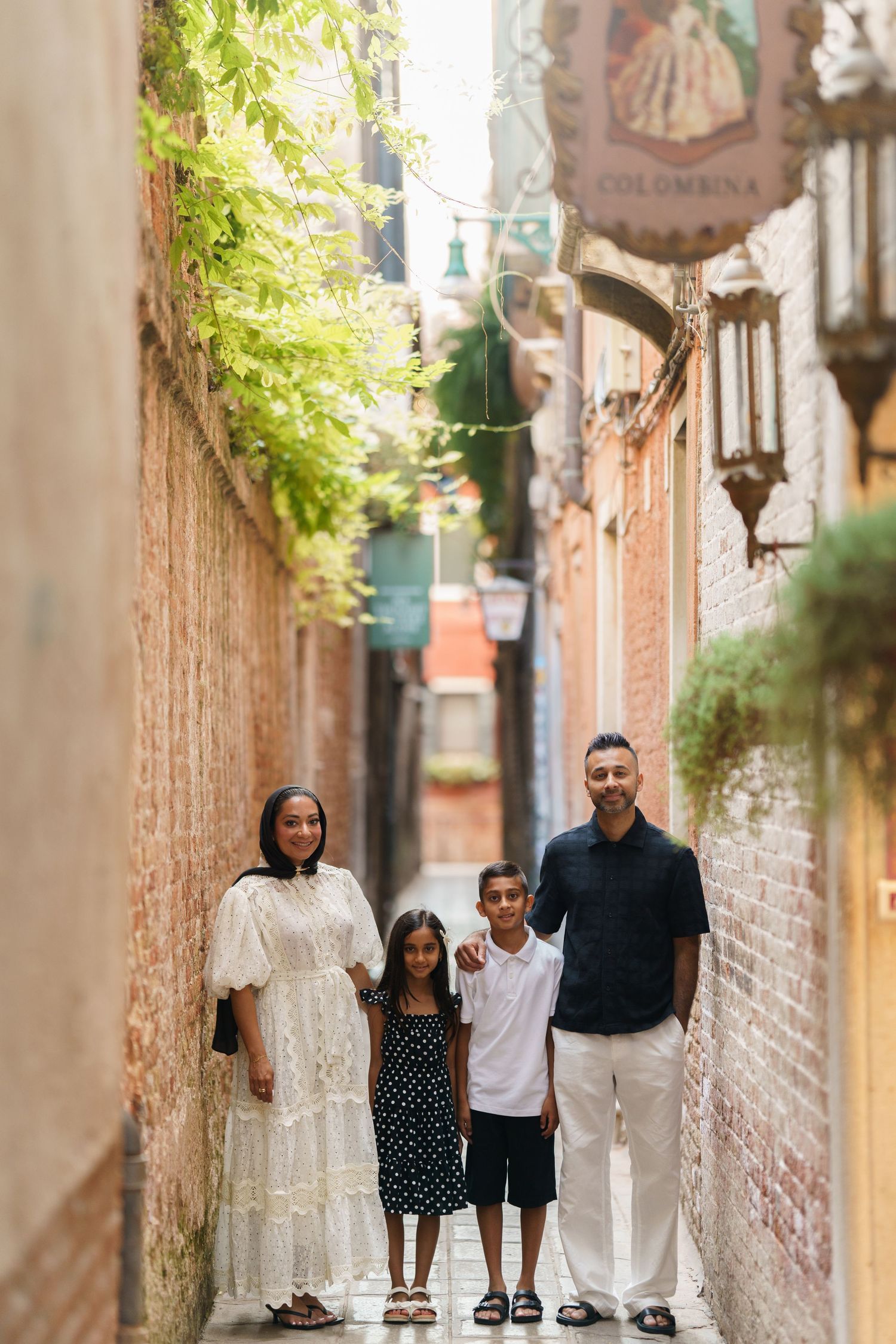 Family of four posing in a charming narrow brick alley in Venice, Italy, with lush greenery overhead.