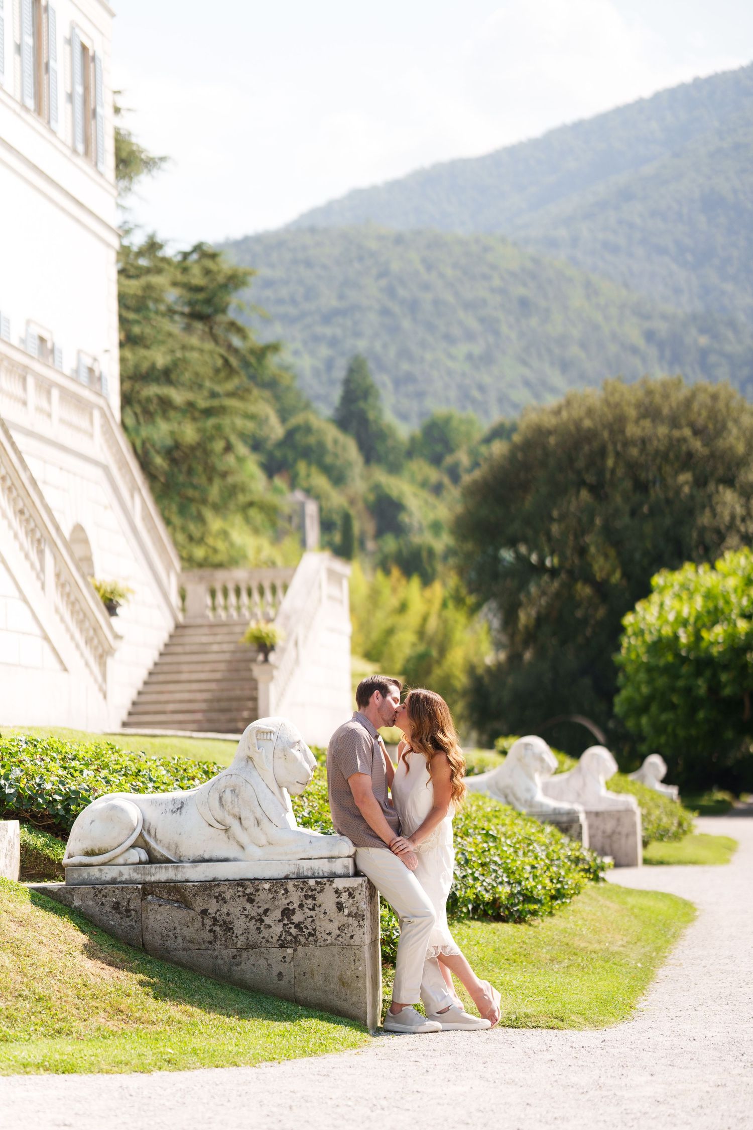 Couple kissing by lion statues at elegant villa with mountain backdrop and manicured gardens.