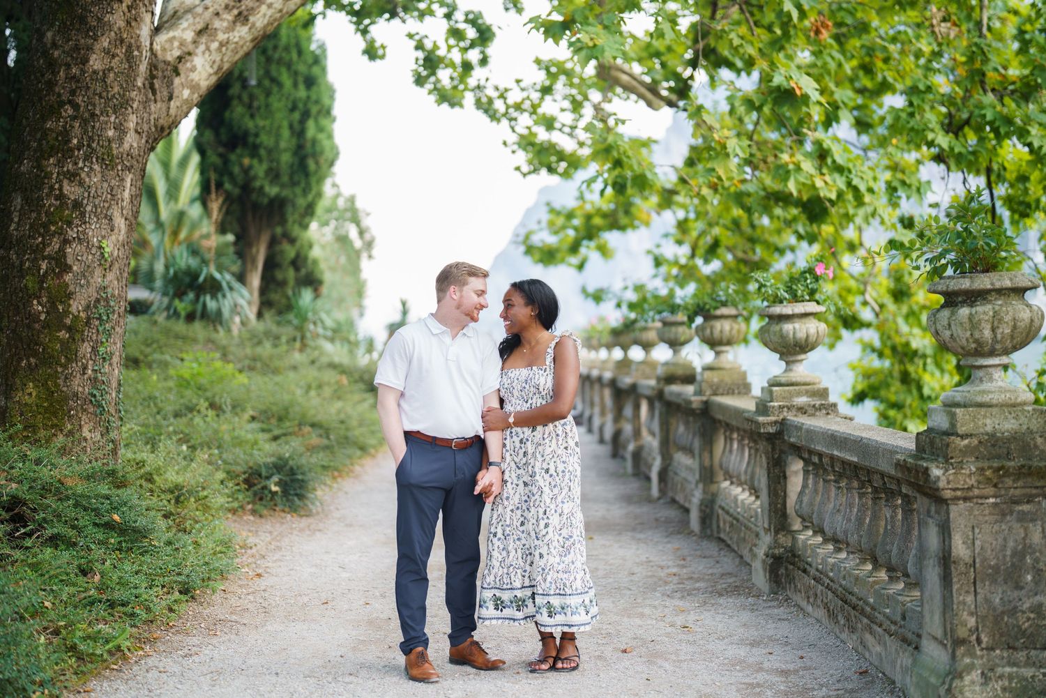 Interracial couple walking and smiling together on a scenic tree-lined path with stone railing.