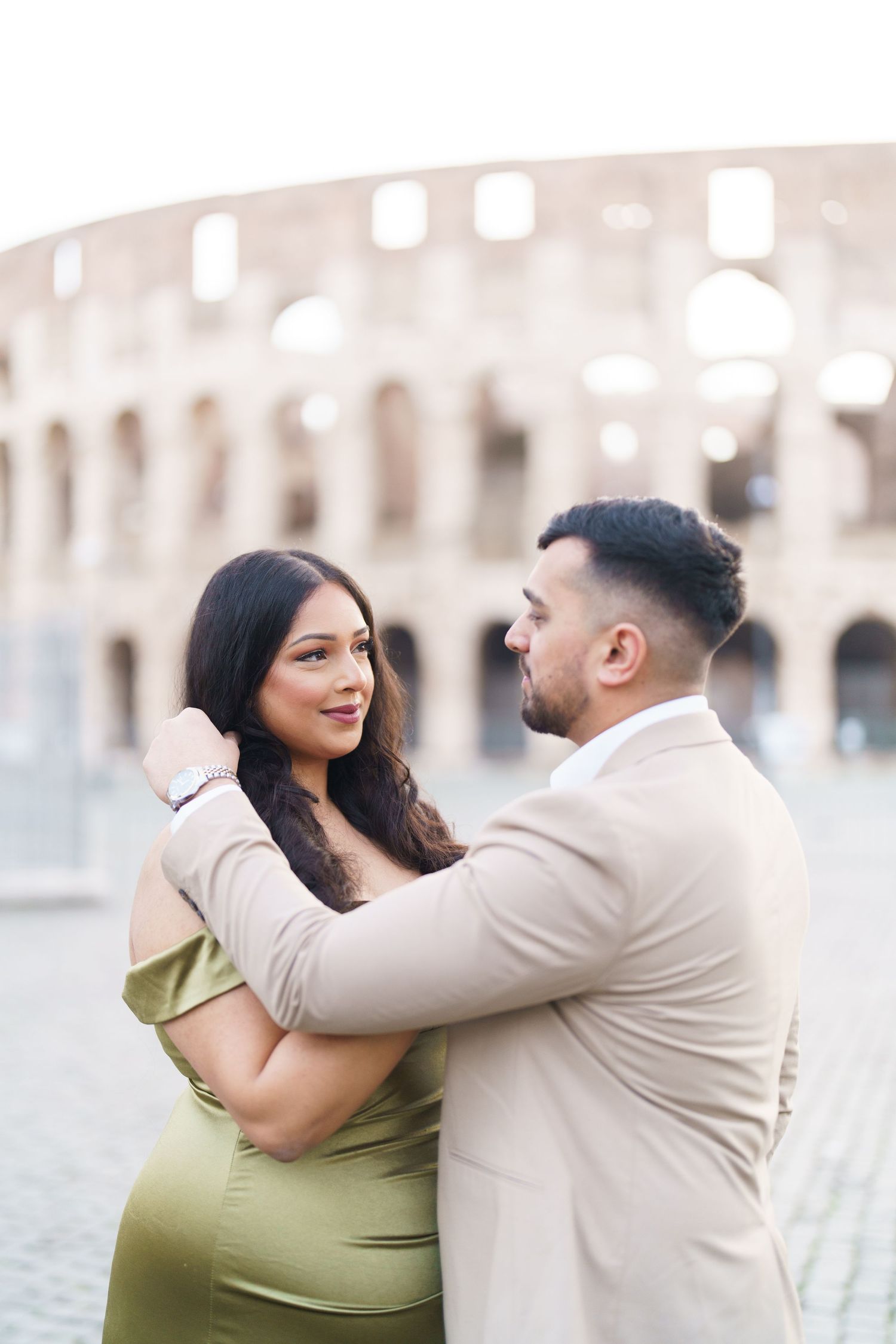 Couple embracing romantically in front of the Colosseum in Rome, woman in green dress and man in beige suit.
