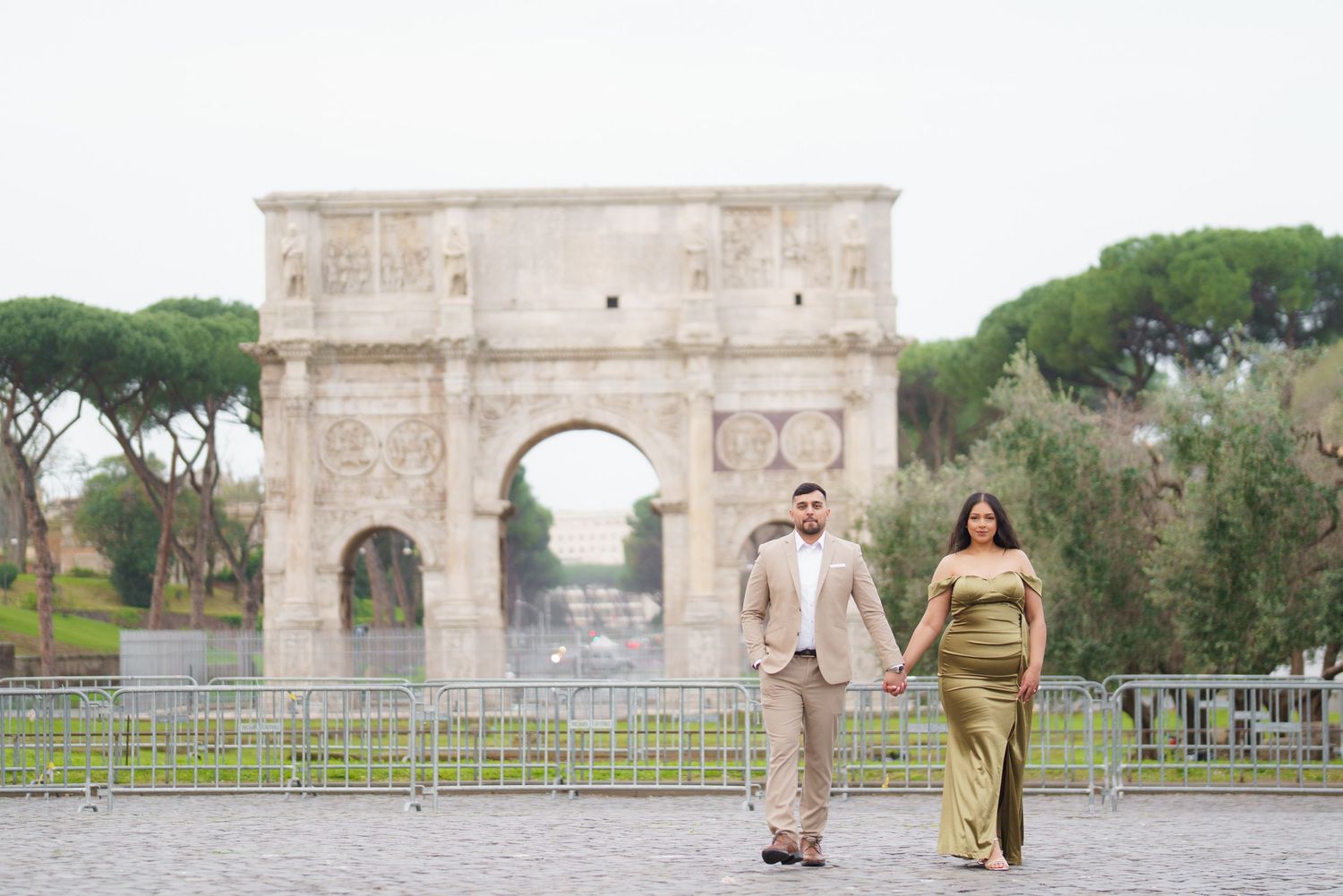 Couple walking hand in hand near the Arch of Constantine in Rome, man in tan suit, woman in gold gown.