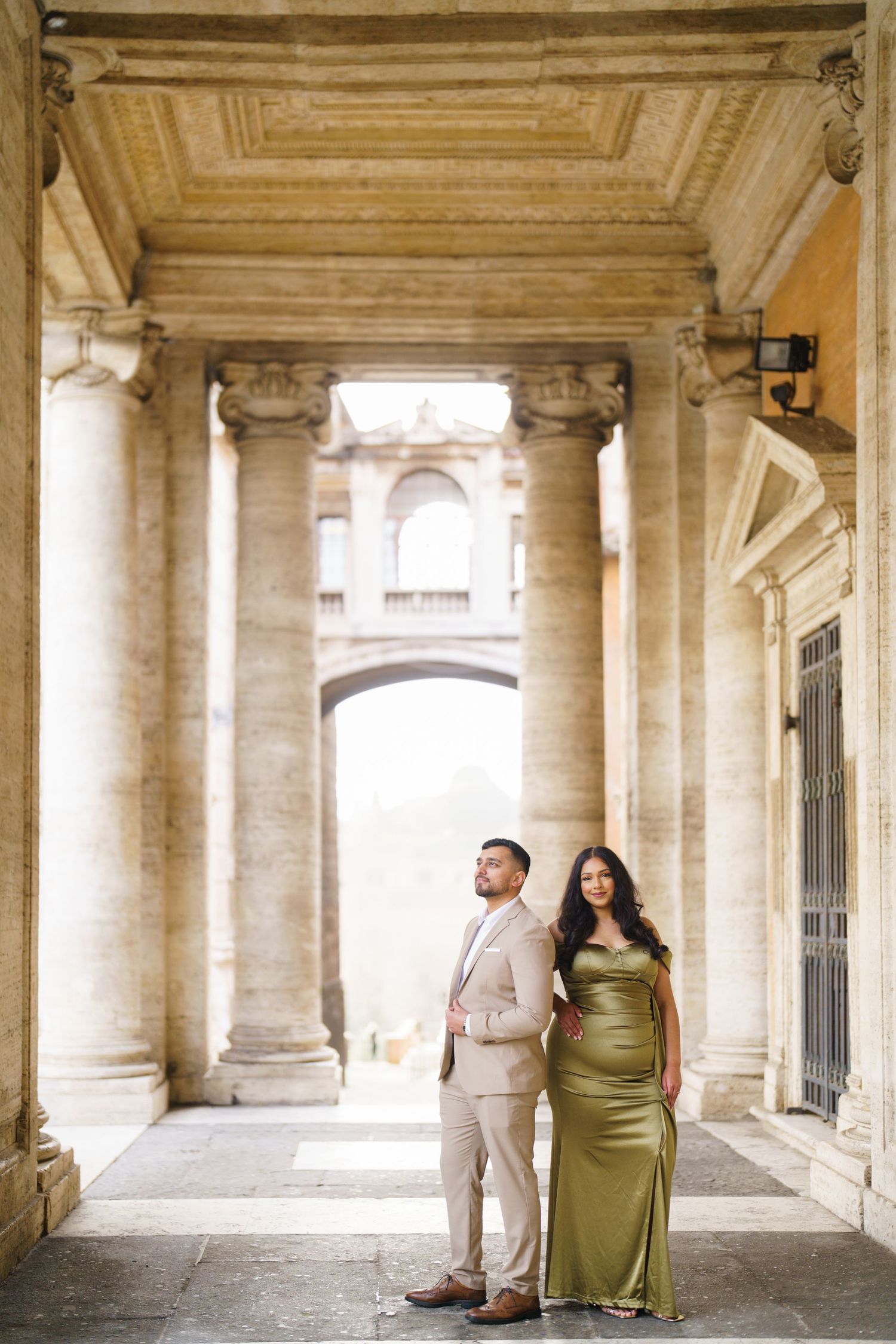 Couple posing under grand stone columns in elegant attire, woman in olive satin gown, man in beige suit, classical architecture.